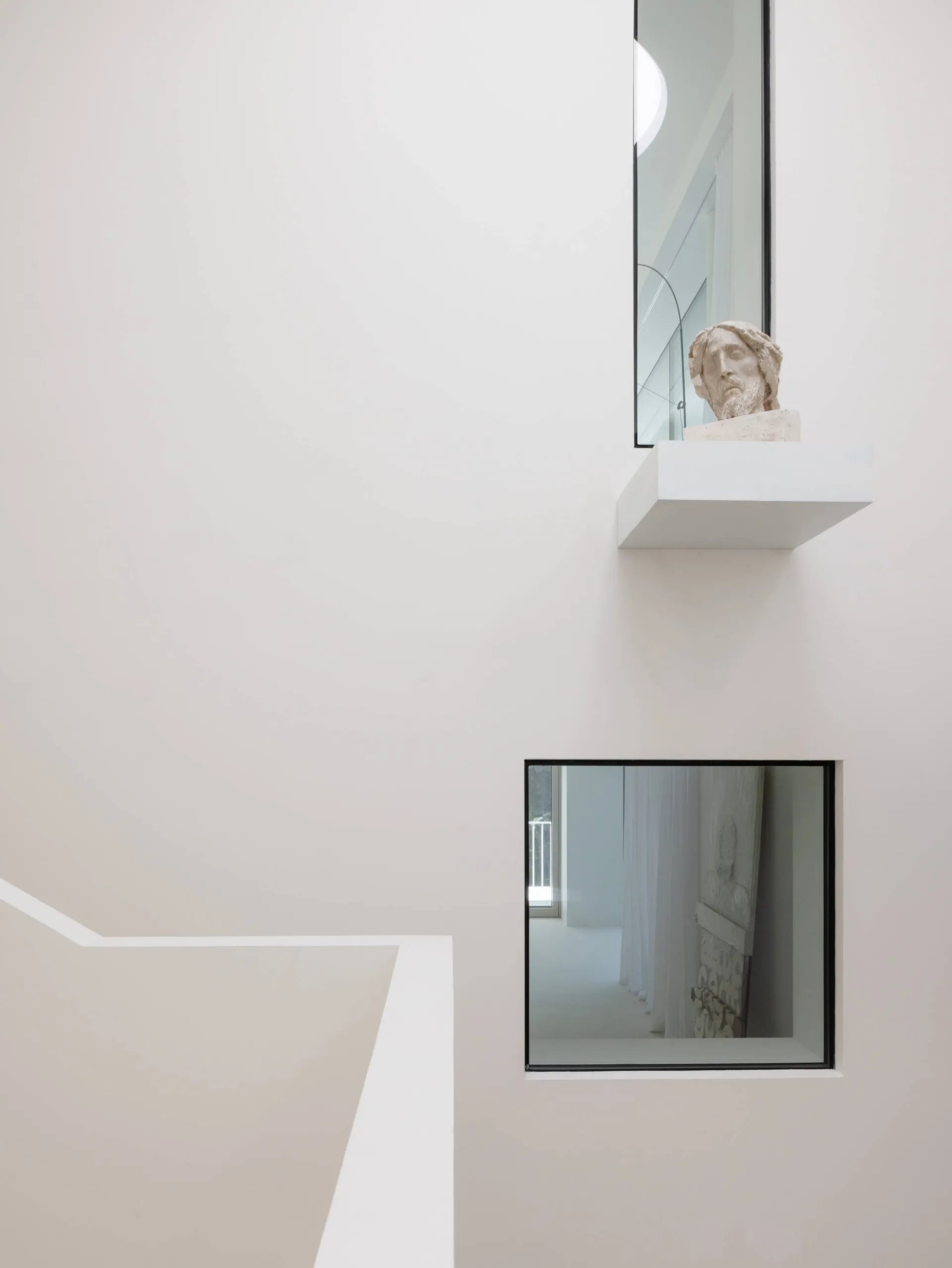 Minimalist white interior featuring a floating concrete shelf with a classical stone bust, viewed from a staircase with square black-framed window below.
