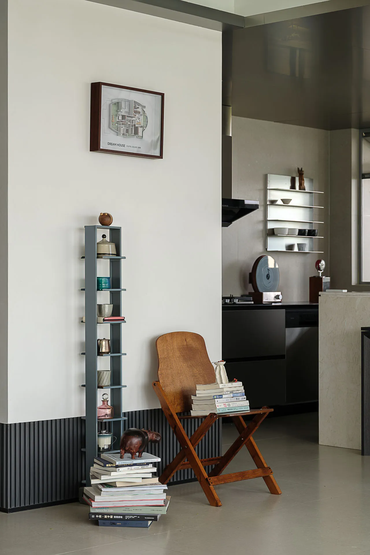 Modern minimalist living room corner featuring a wooden folding chair used as a side table, a tall blue open shelving unit, stacks of books, and a framed architectural floor plan on the wall.