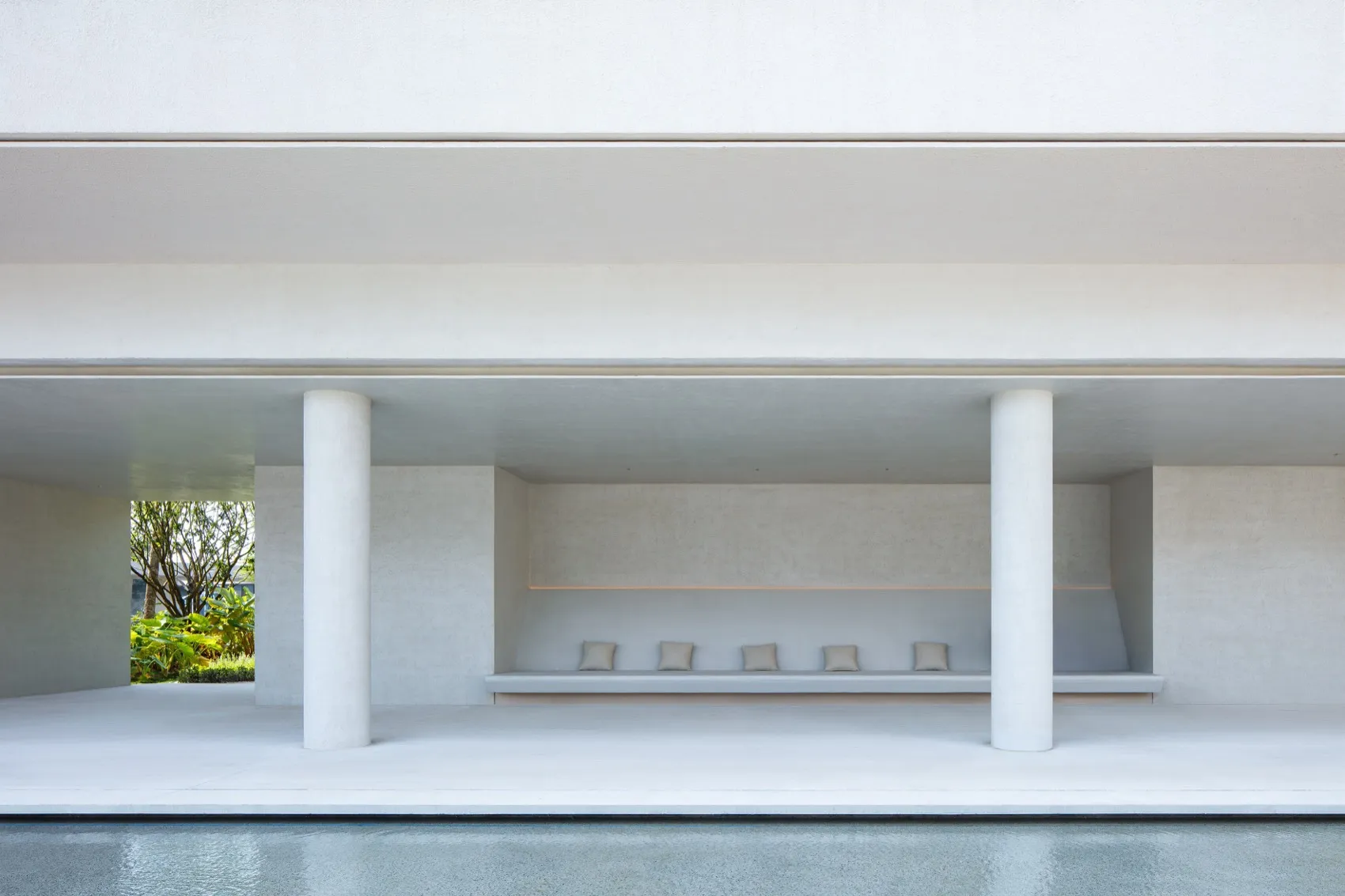 Modern minimalist concrete outdoor pavilion with cylindrical columns, built-in bench with five cushions, and uplighting, opening to a lush garden.