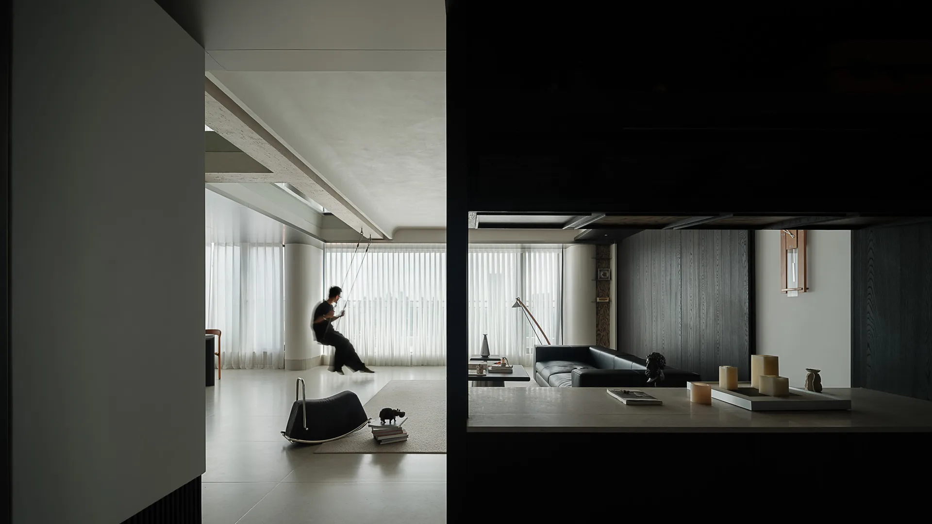 Modern minimalist living room featuring a hanging indoor swing, micro-cement walls and ceiling, dark wood paneling, and a black leather sofa under soft natural light.