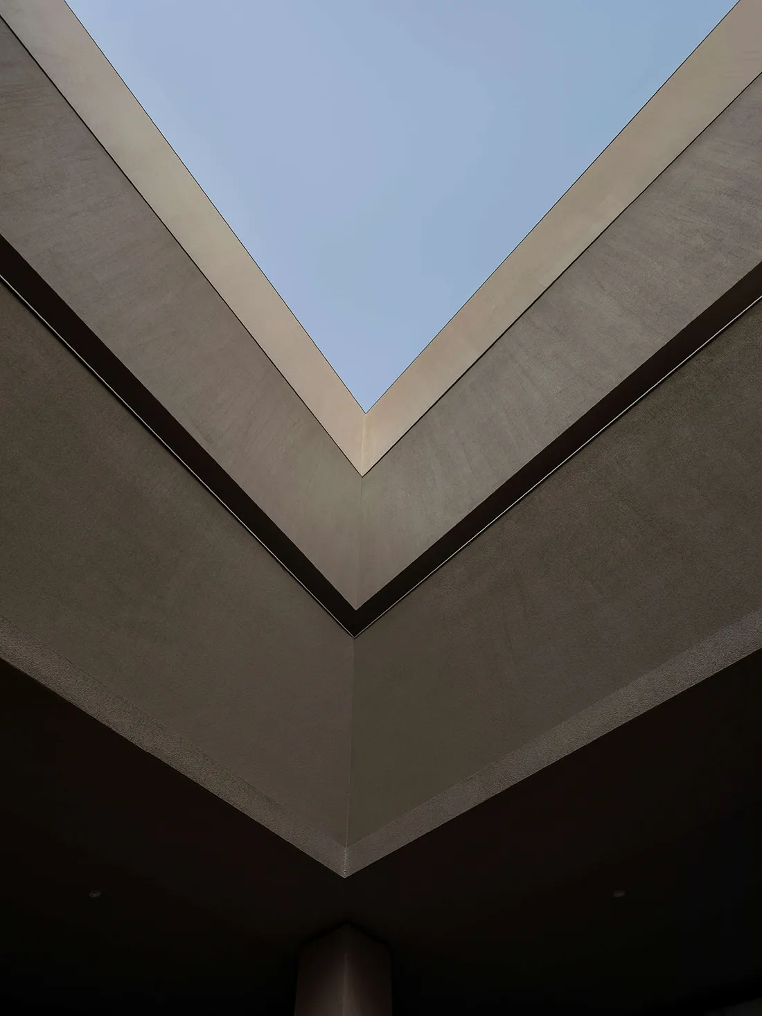 Looking straight up at a modern V-shaped concrete ceiling with a large rectangular skylight opening to blue sky.