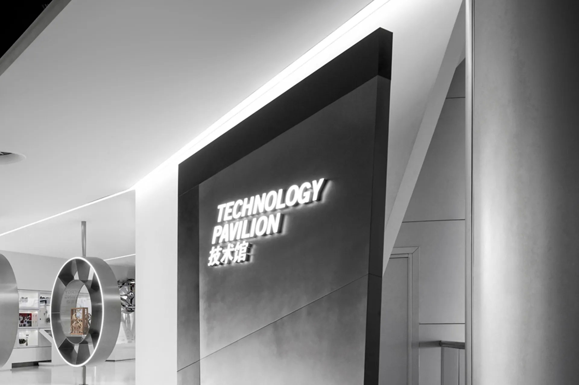 Monochrome interior of a technology pavilion featuring an illuminated 3D sign reading TECHNOLOGY PAVILION with Chinese characters, set against minimalist white walls and metallic circular structures.