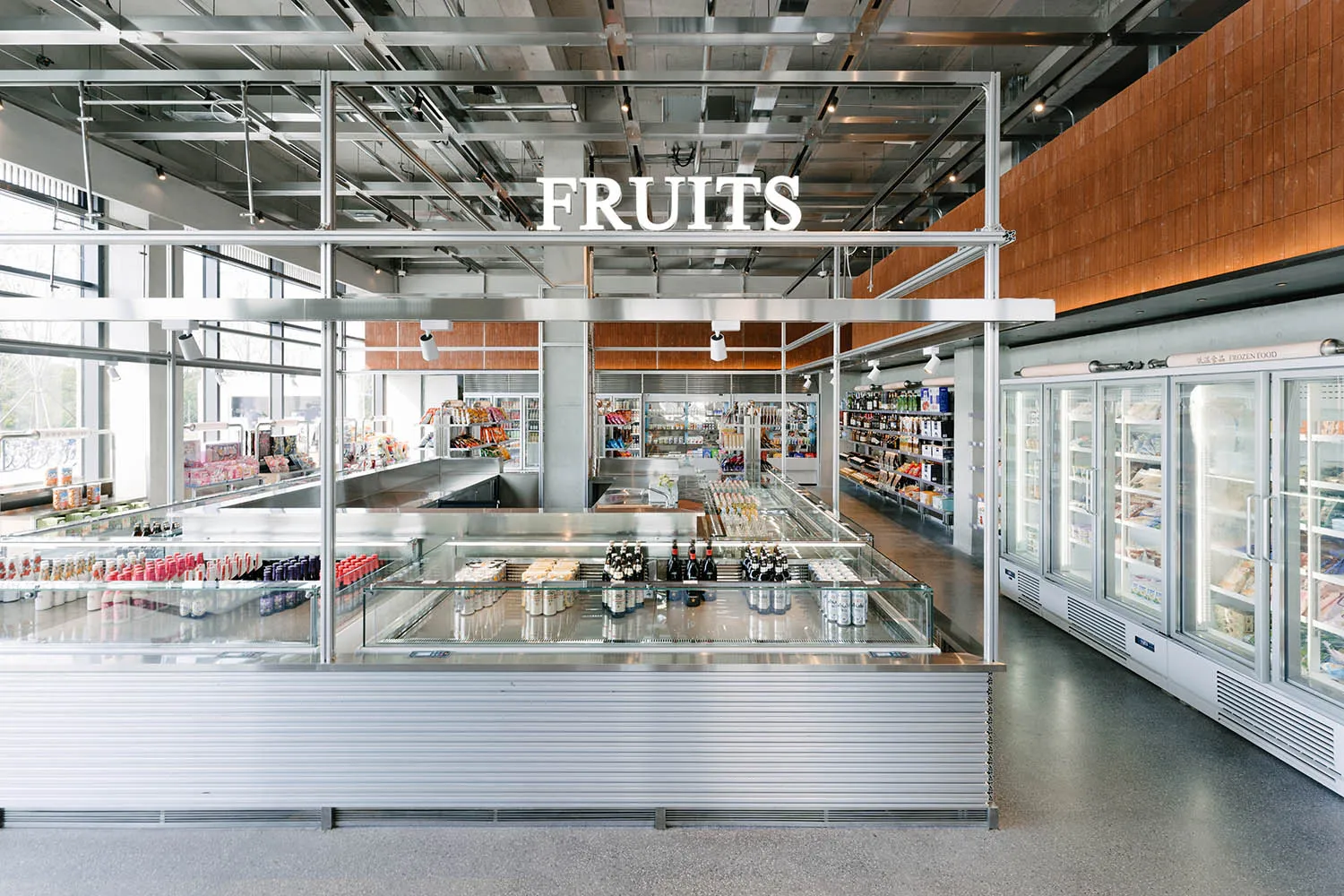 Wide view of a modern industrial-style grocery store featuring a large stainless steel central display island, glass refrigerated cases, concrete floors, and exposed ceiling infrastructure with a prominent FRUITS sign.