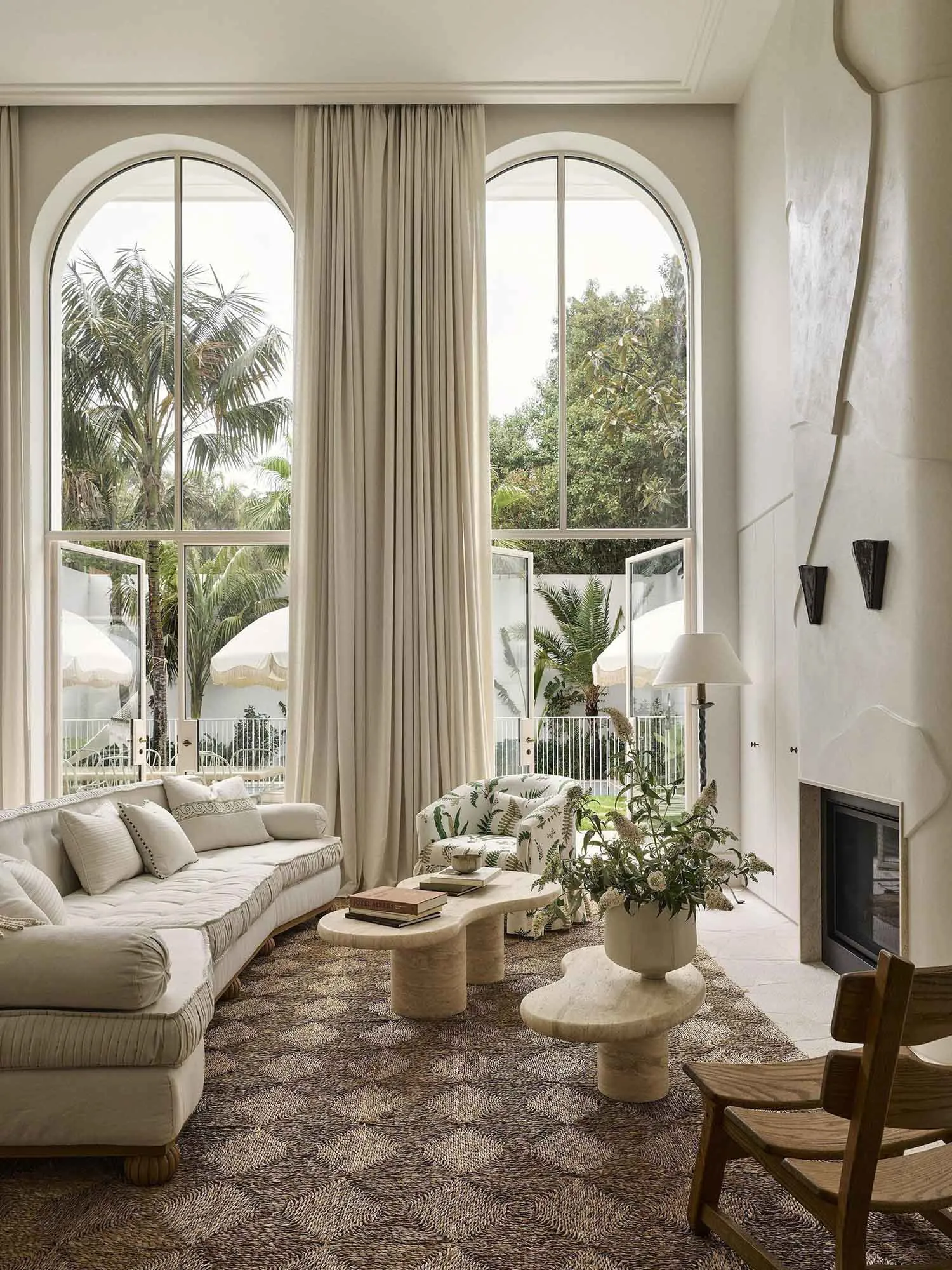 Bright living room featuring a curved white sofa, patterned rug, organic travertine coffee tables, a tropical print armchair, and large arched windows overlooking a palm-filled garden.
