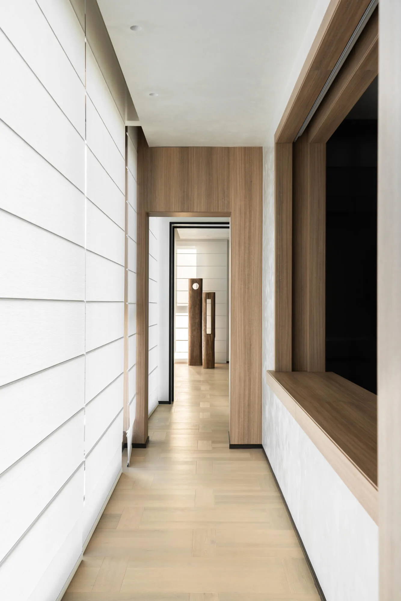 Modern minimalist hallway featuring white horizontal wall paneling, light oak wood accents, built-in window bench, and sculptural wooden pillars visible in the distance.