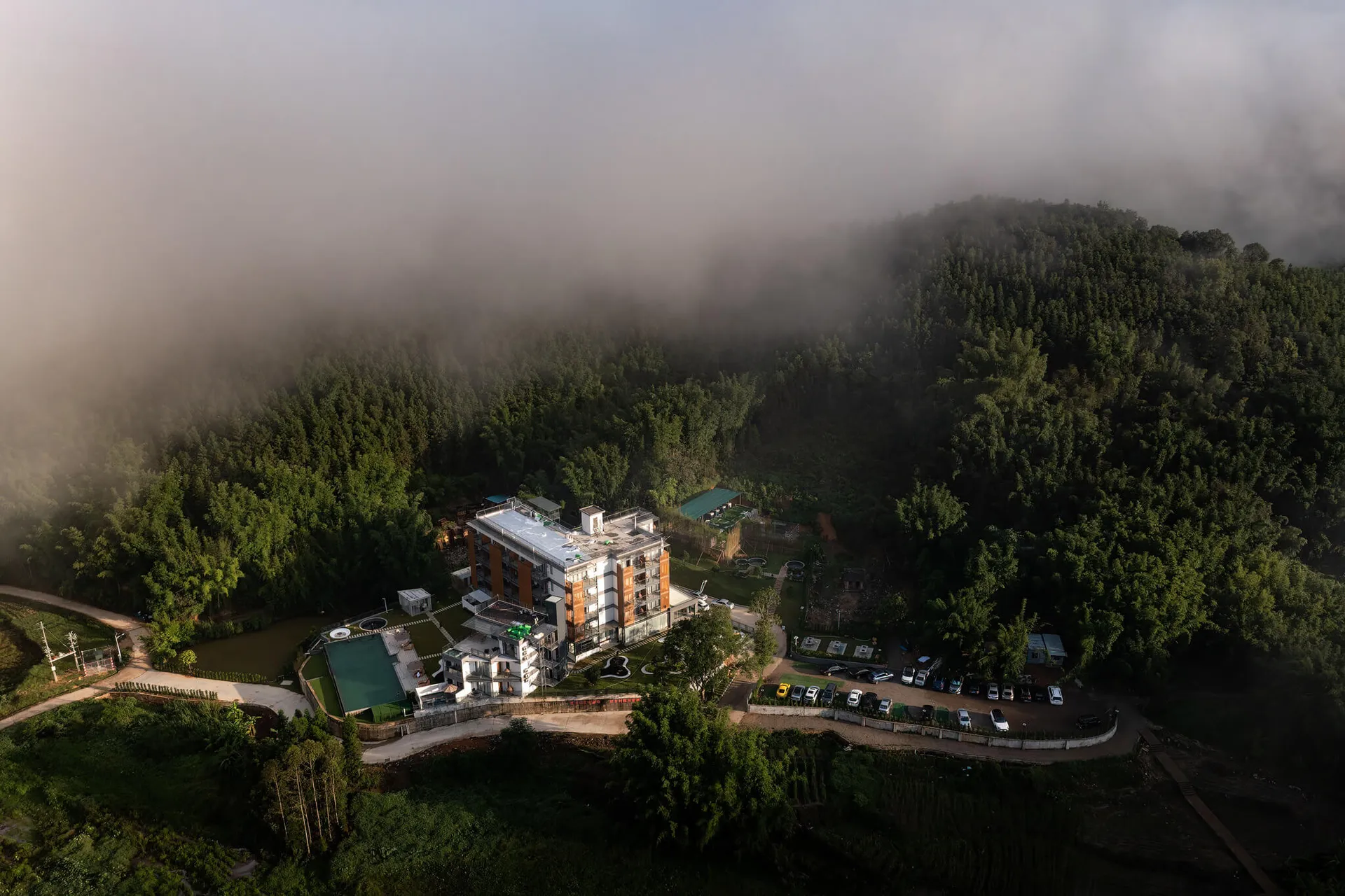 航拍照片顯示一座結合混凝土與玻璃立面的多層現代度假村,坐落於茂密森林與濃厚白霧的山區,周圍可見游泳池與停車場。