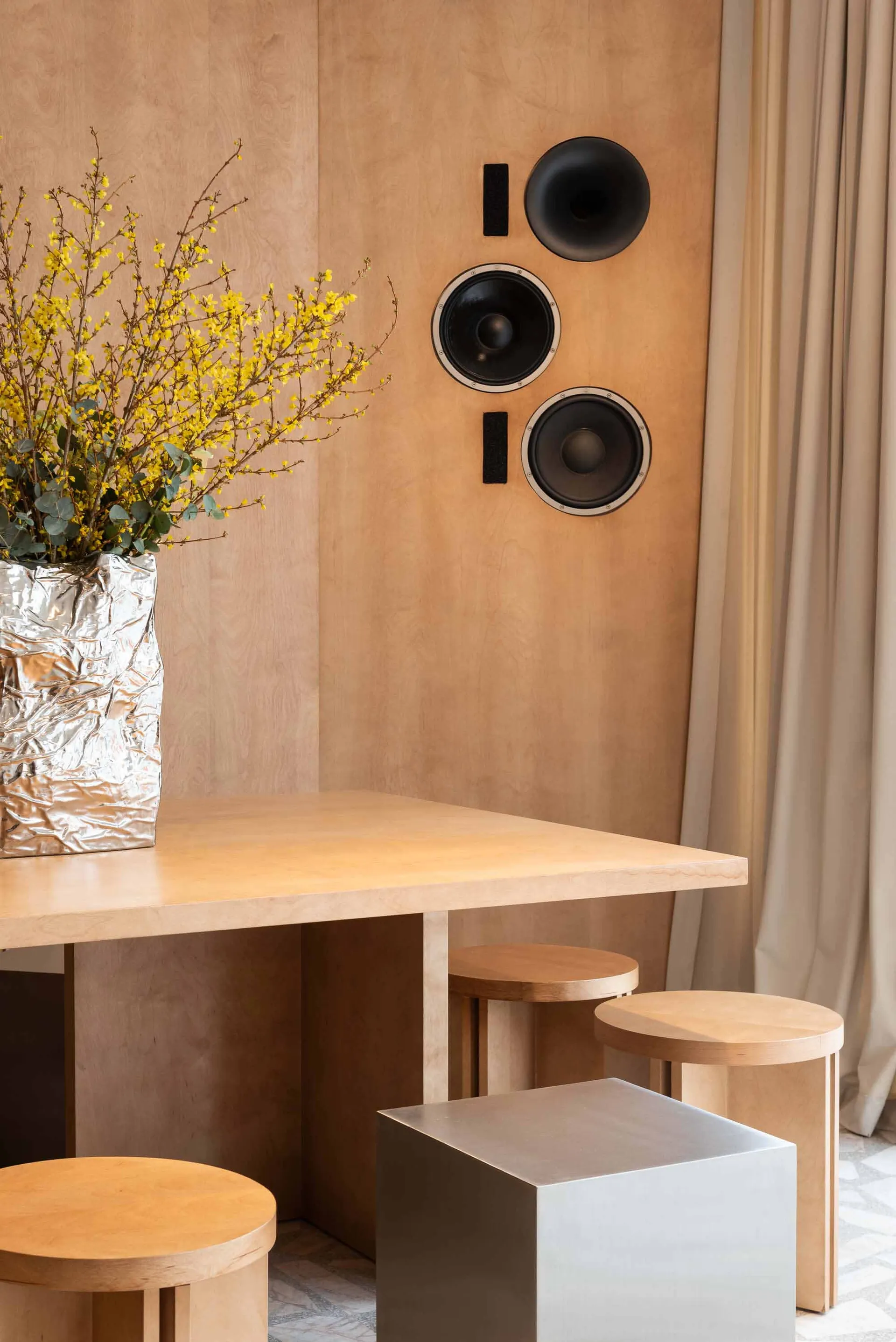 Modern dining area featuring a light birch plywood wall with mounted black speakers, a matching birch table, cylindrical wooden stools, a metallic cube stool, and a large silver foil vase filled with yellow forsythia branches.