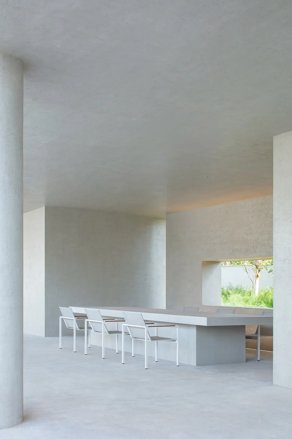 Minimalist concrete dining room featuring a long built-in concrete table, white metal-framed chairs, and a large horizontal window overlooking a green garden.