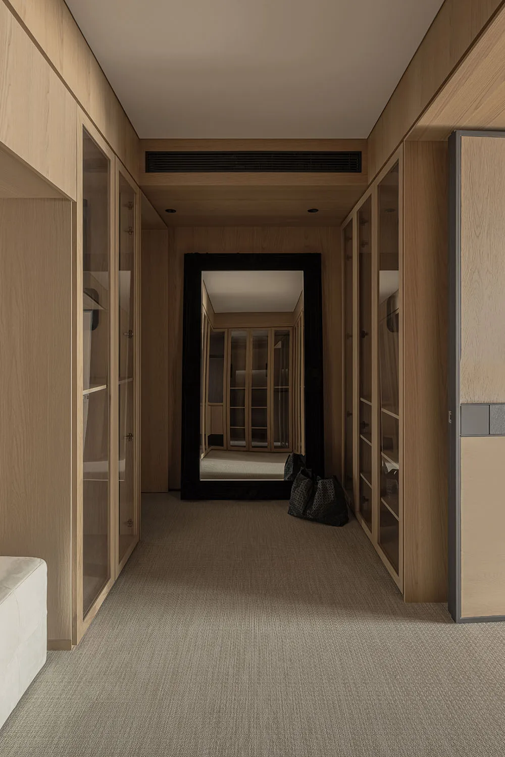 Narrow walk-in closet featuring light oak wood cabinetry with glass doors, beige carpet flooring, and a large black-framed mirror reflecting another closet space.