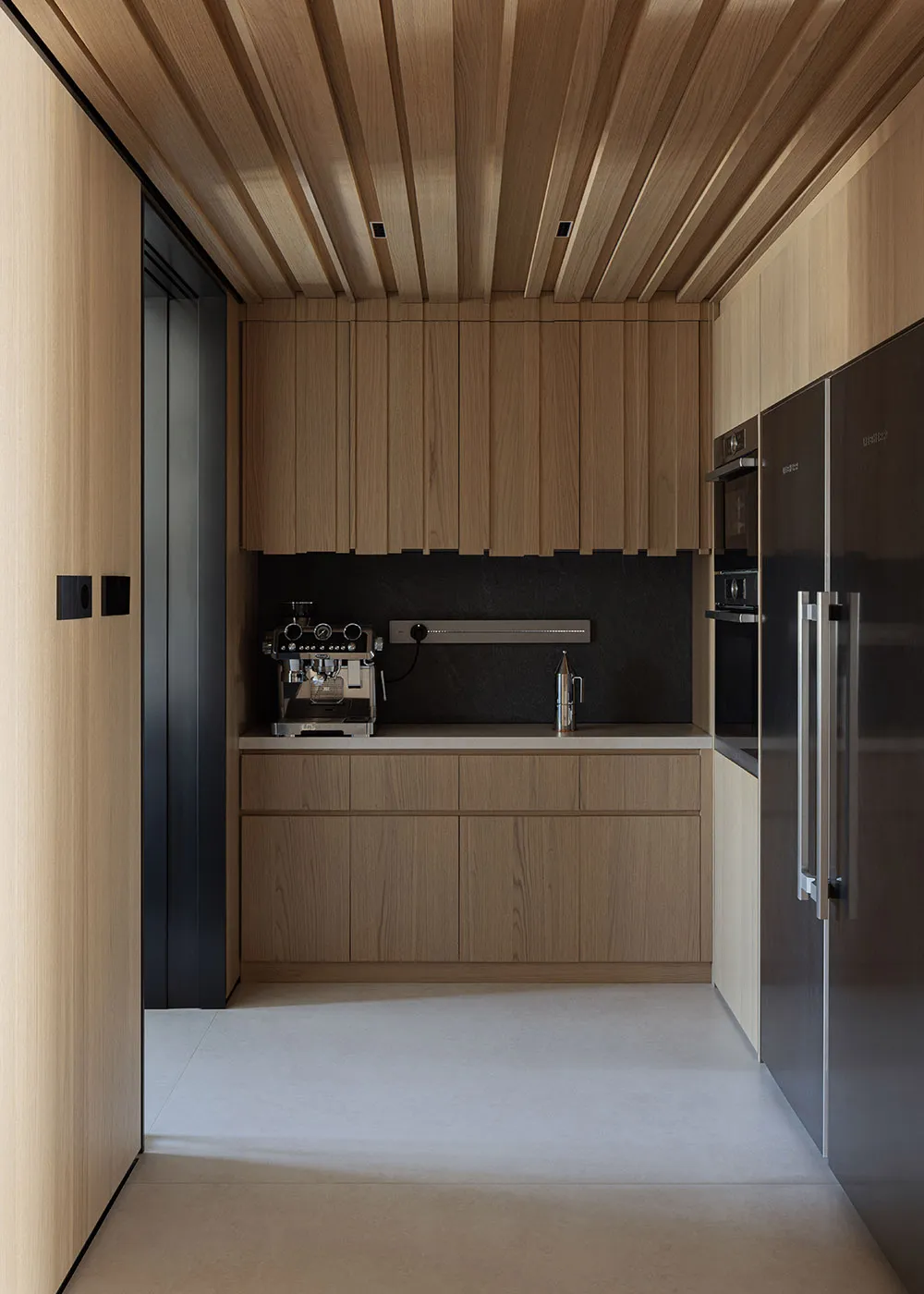 Modern narrow butler pantry with light oak wood cabinetry, black stone accent wall, wood slat ceiling, built-in coffee station, and stainless steel refrigerator.