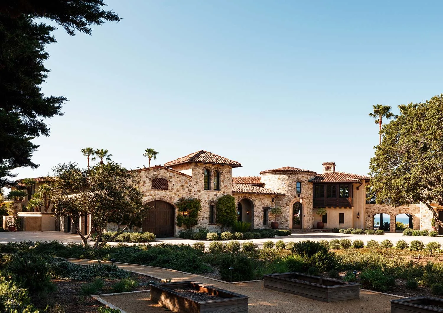 Expansive Tuscan-style stone villa with terracotta roof, arched windows, lush Mediterranean garden and distant ocean view under clear blue sky.