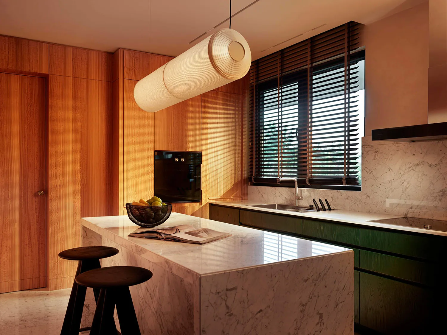 Modern kitchen featuring a white marble island with dark wood stools, warm vertical wood paneling, forest green lower cabinets, and a long cylindrical fabric pendant light.