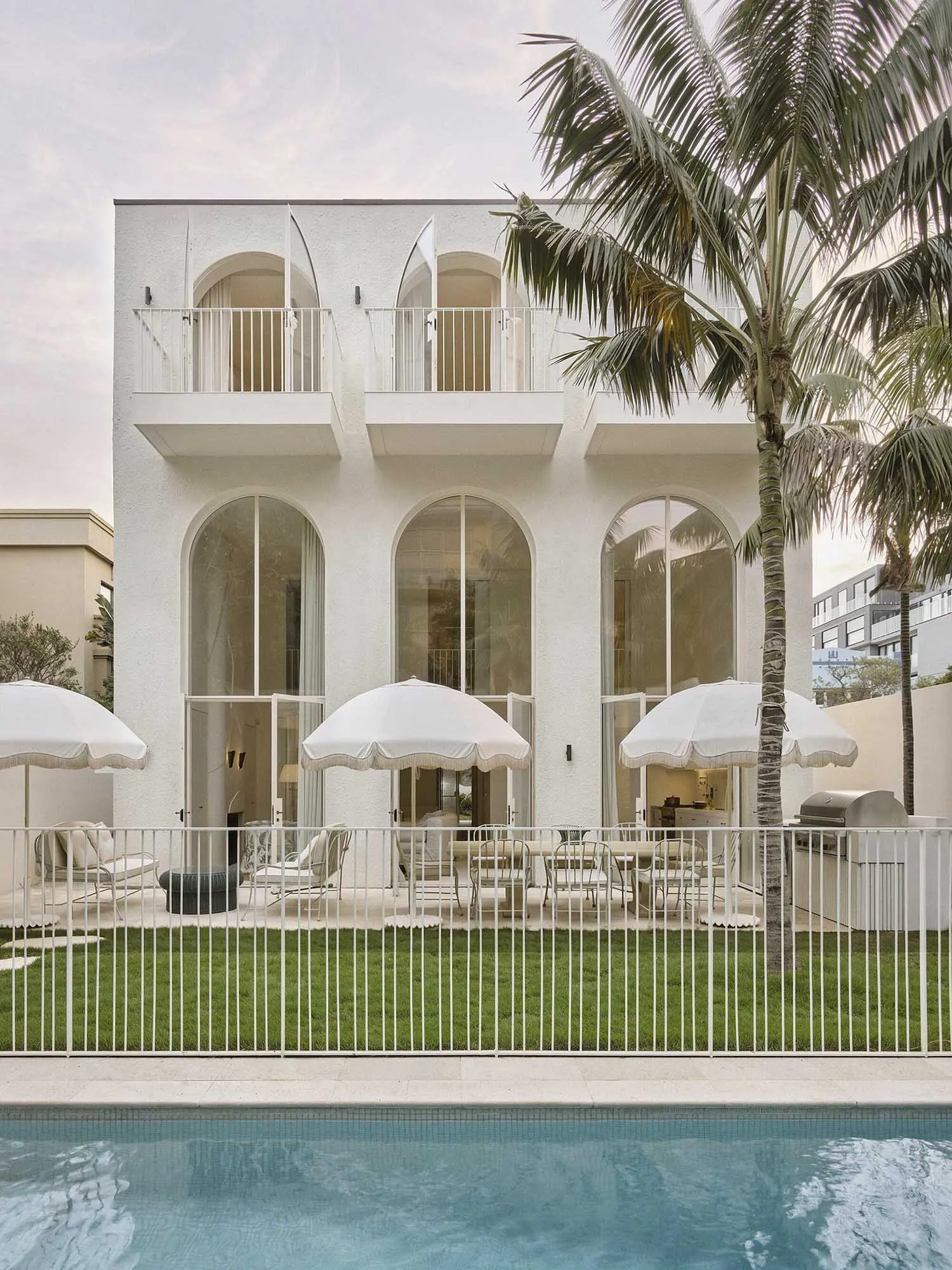Modern white stucco villa with large arched windows and doors opening to a lush green lawn, outdoor dining area with white umbrellas, and a swimming pool in the foreground under palm trees.