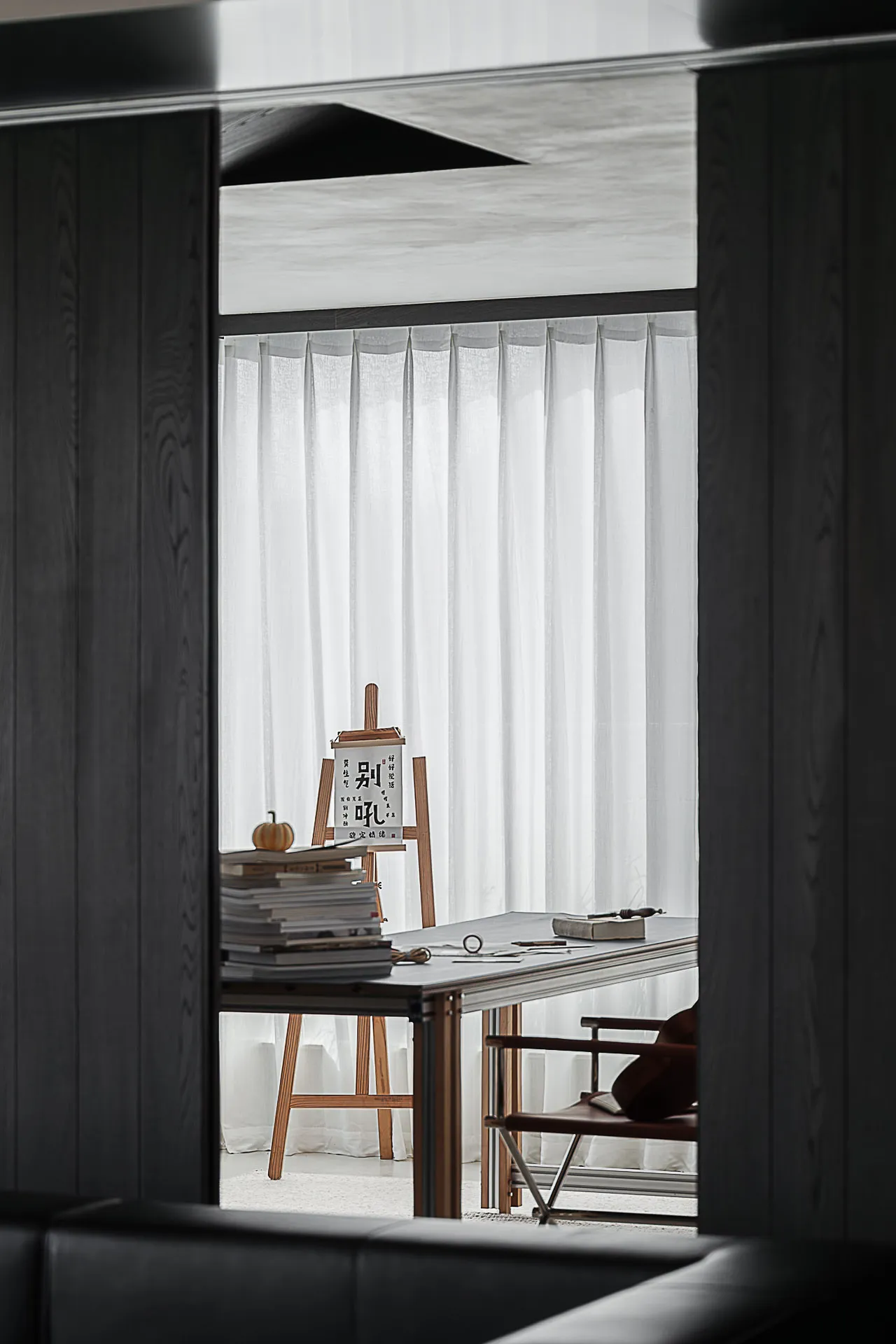 View through a dark wood partition into a bright minimalist home office featuring a wooden easel with calligraphy, stacked books, a simple desk, and a leather chair by the window with sheer white curtains.