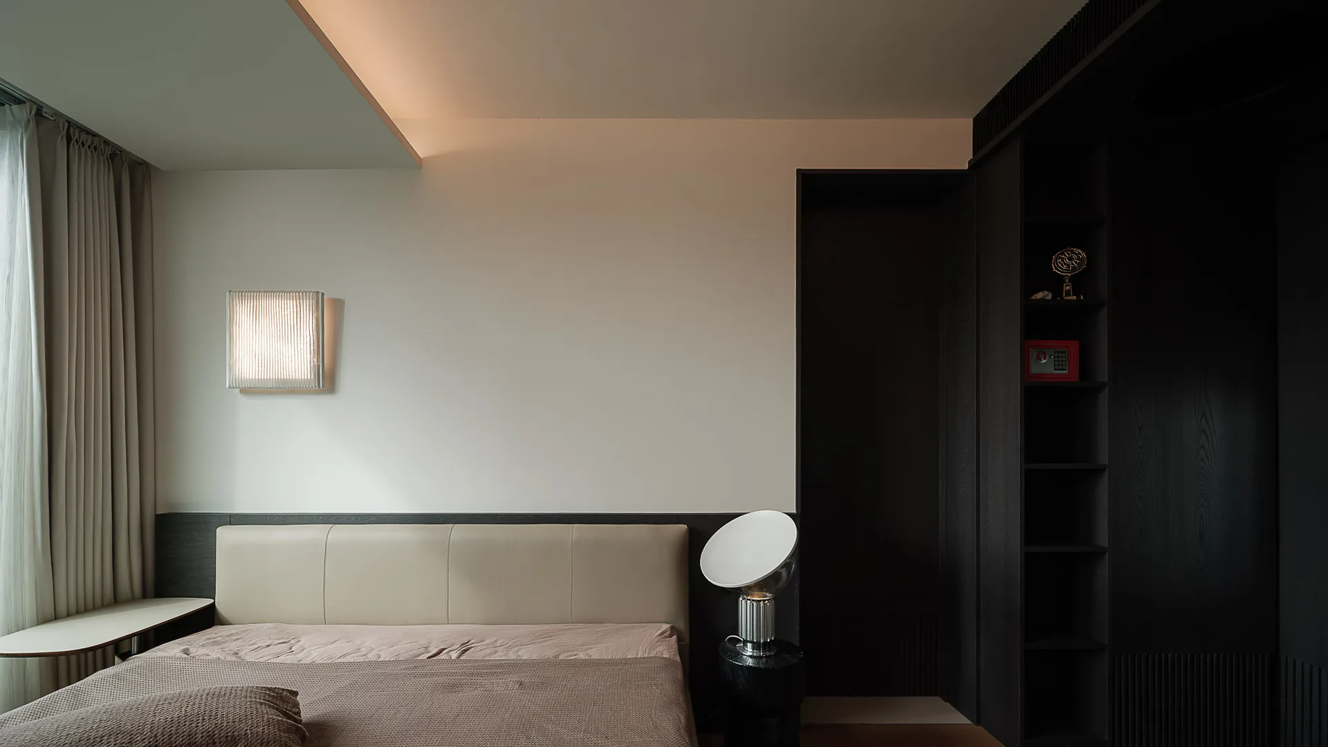 Modern minimalist bedroom with beige leather headboard, dark wood built-in cabinetry, wall-mounted light fixture, and spherical table lamp.