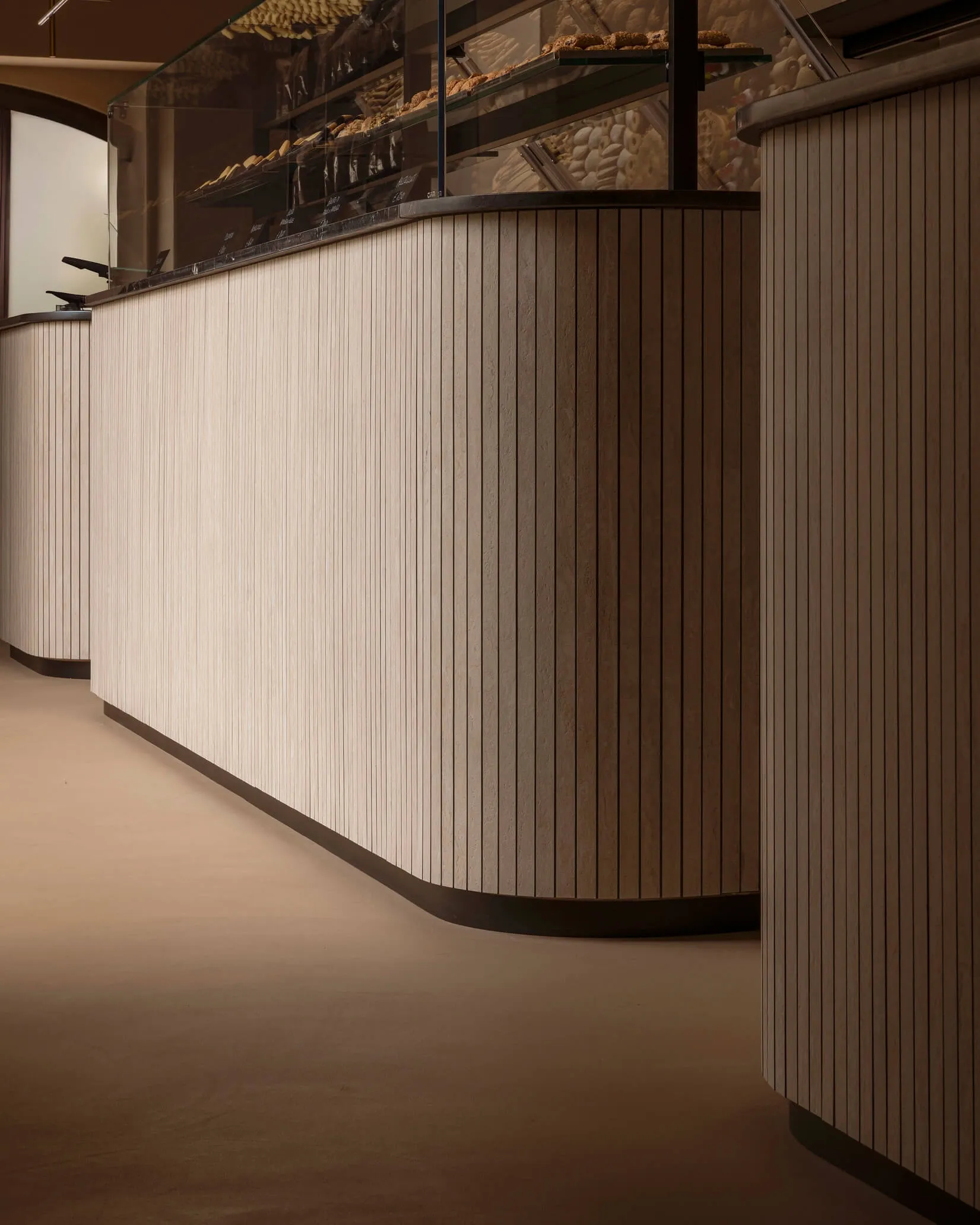 Modern bakery service counter with curved fluted wood paneling, black stone countertop, and glass pastry display cases filled with baked goods.