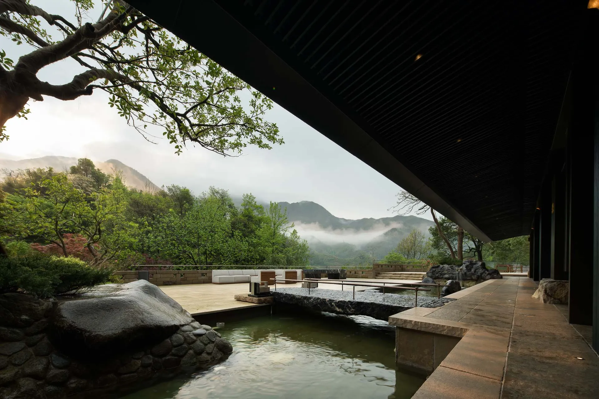 Modern Japanese-style garden with stone-lined pond, wooden deck, white outdoor sofas, and lush greenery overlooking misty mountains.