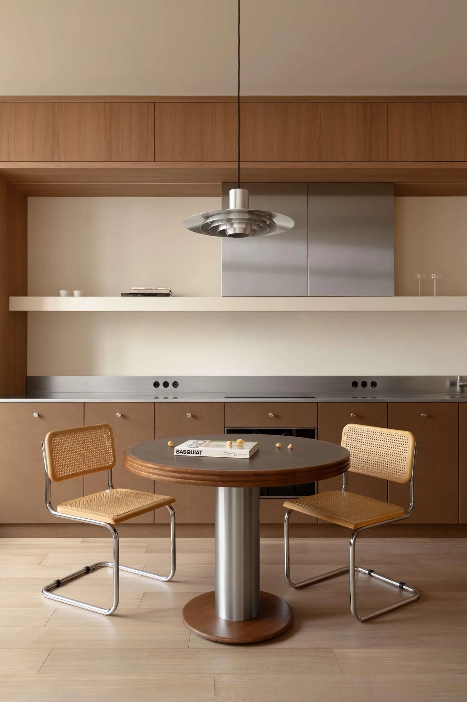 Modern minimalist dining area with a round walnut table, two cane and chrome chairs, stainless steel kitchen counter, and wooden upper cabinets under a sculptural metallic pendant lamp.