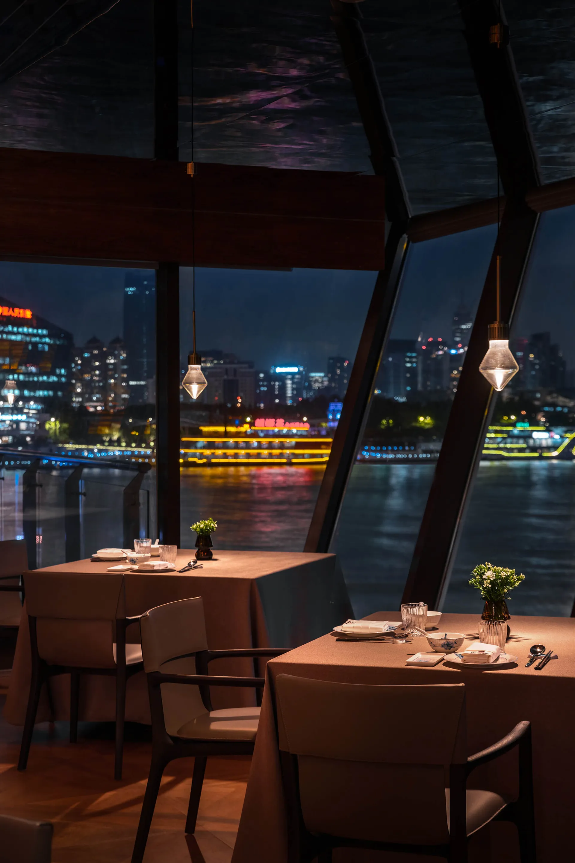 Elegant modern restaurant interior at night featuring two set dining tables with beige tablecloths and leather chairs, hanging glass pendant lights, dark wooden ceiling beams, and a stunning nighttime view of a river and illuminated city skyline through floor-to-ceiling angled windows.