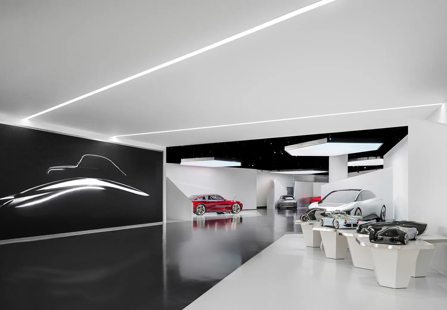 現代汽車展覽空間,展示多輛概念車置於白色基座上,大型黑色牆面有銀色汽車剪影,搭配拋光深色混凝土地板與戲劇性線性LED燈光。
