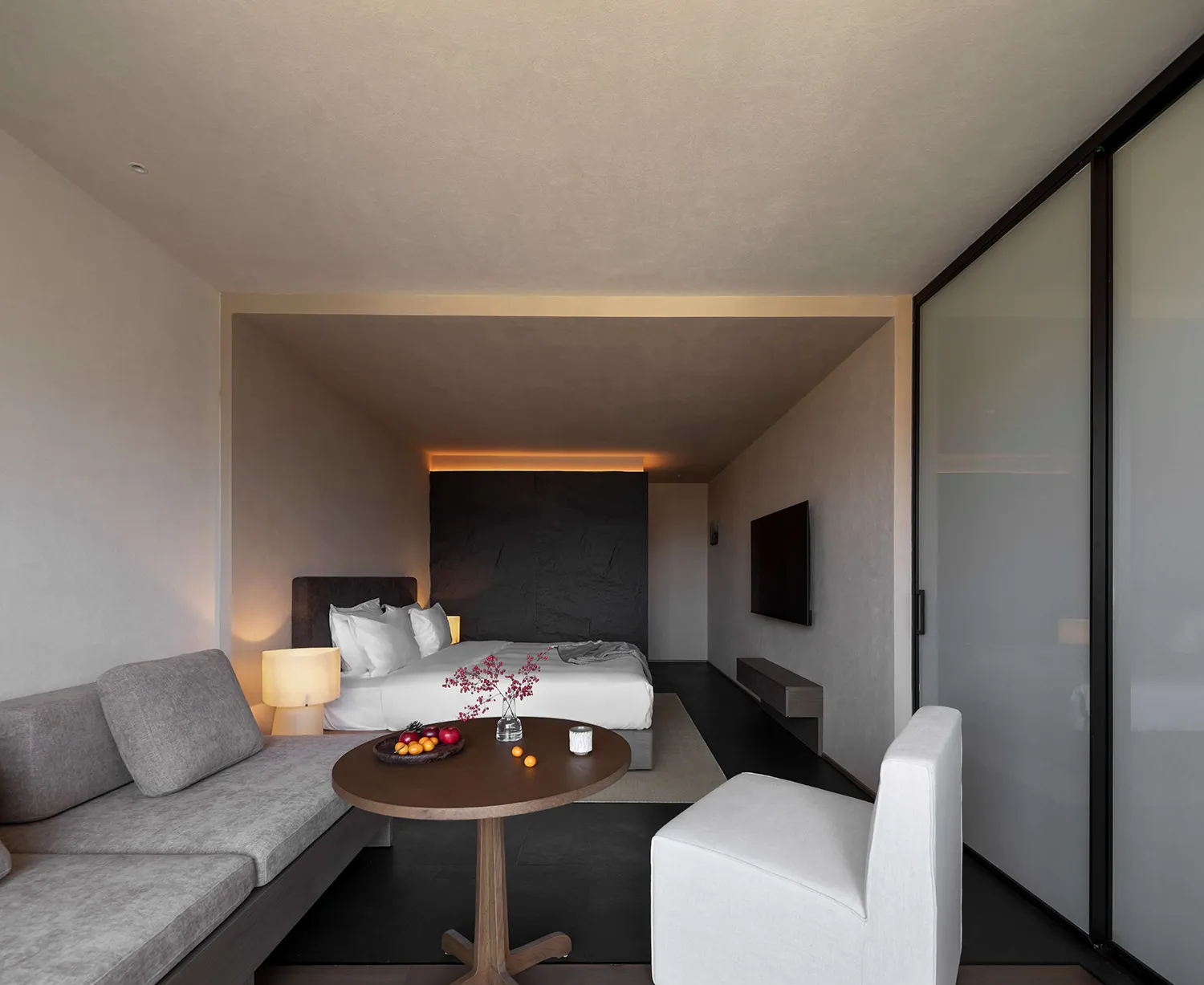 Modern minimalist bedroom featuring a dark textured stone accent wall, white bedding, a gray built-in sofa, round wooden coffee table, and a white armchair.