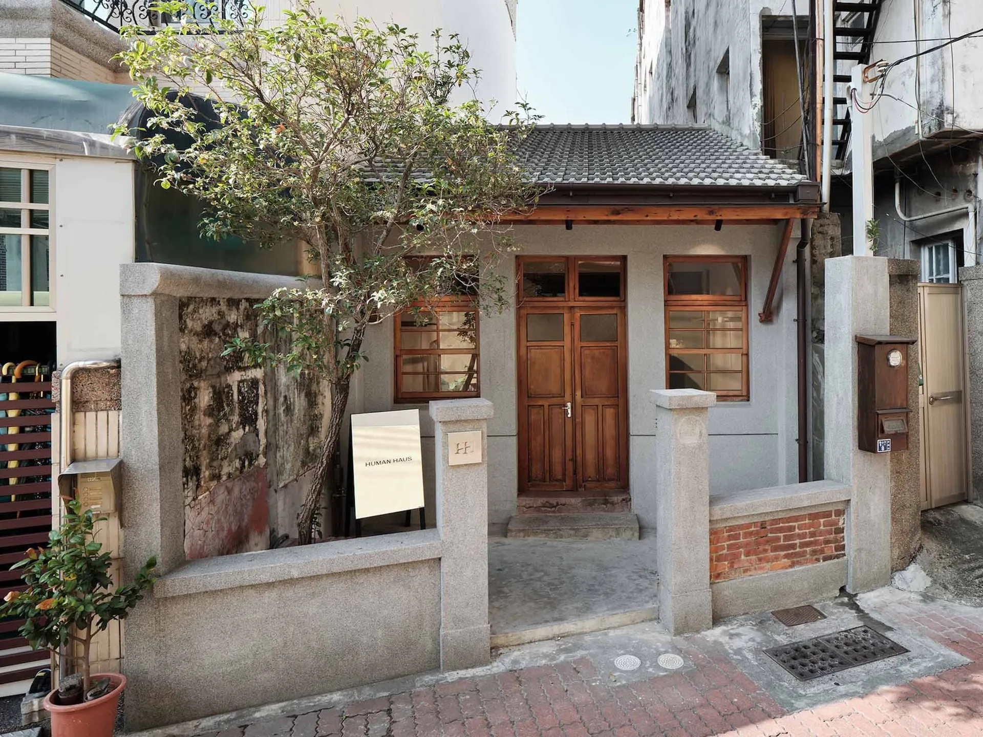 Street view of a renovated old house entrance featuring a grey concrete wall, wooden double door, small tree, and brick pavement in a narrow alley.