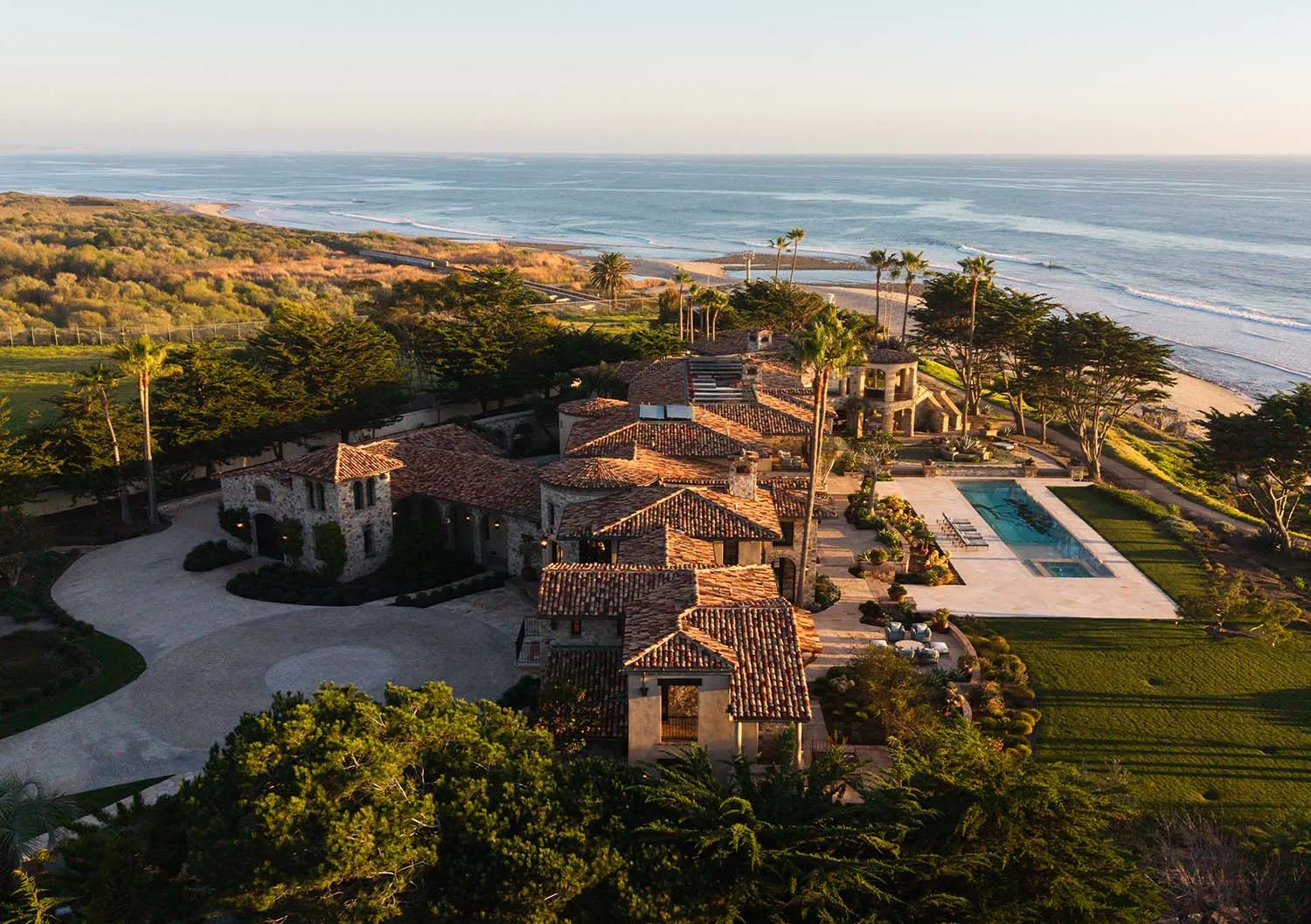 Aerial view of a luxurious Mediterranean-style stone villa with terracotta tile roofs, swimming pool, and manicured gardens overlooking the Pacific Ocean at sunset.