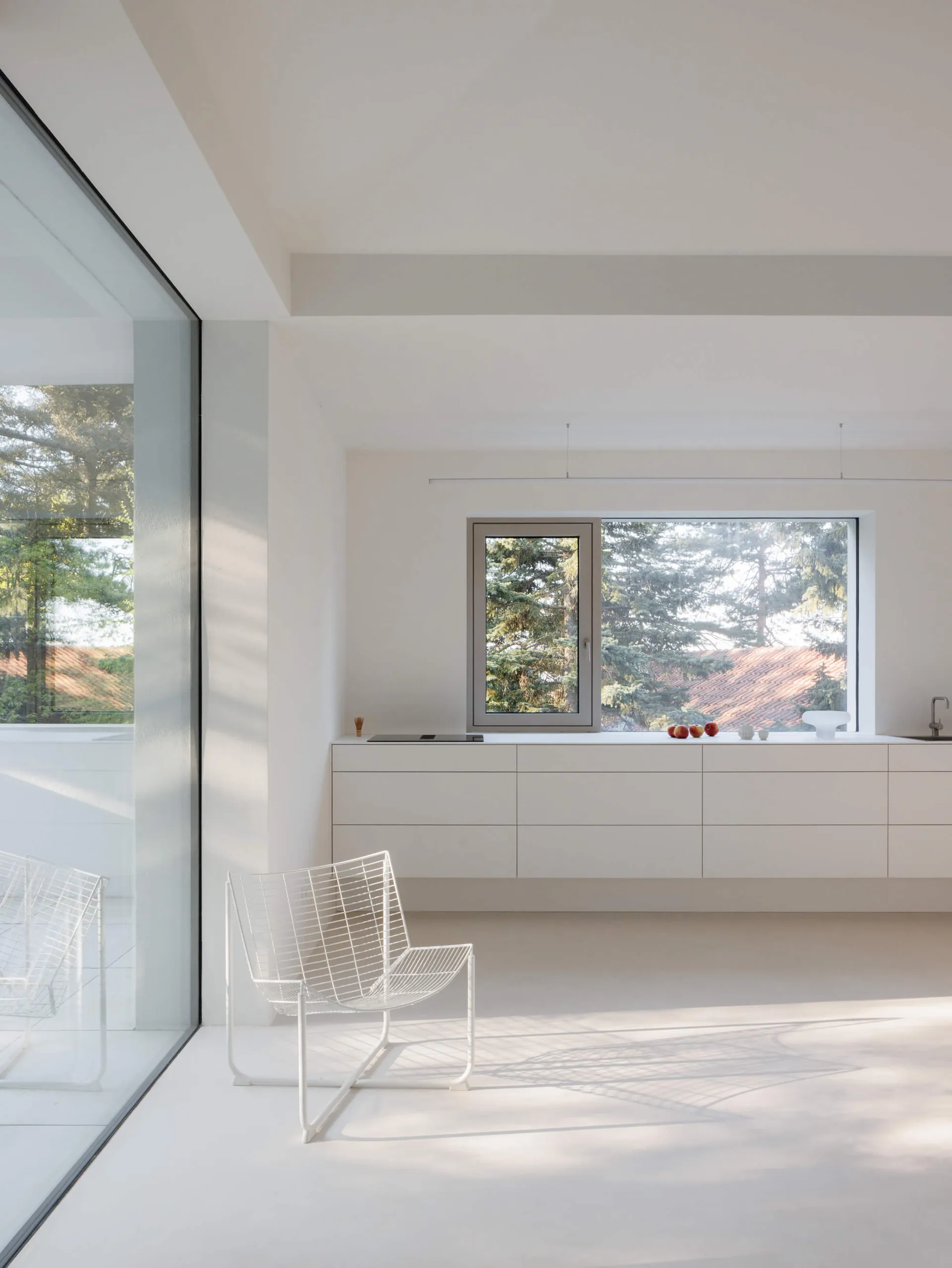 A bright, minimalist living room with white walls, a white wireframe chair, floating white cabinetry, and large windows overlooking green trees and a tiled roof.