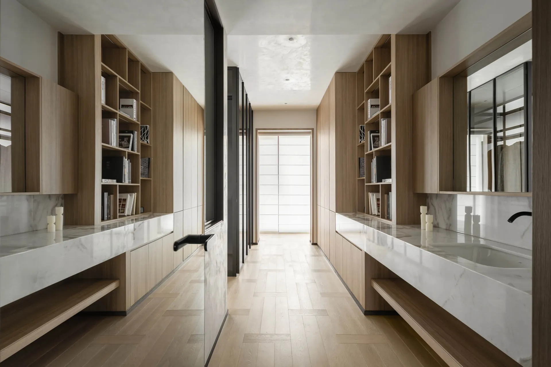 Modern minimalist walk-in closet with light oak built-in shelving, white marble countertops, and a central dressing aisle leading to a bright window.