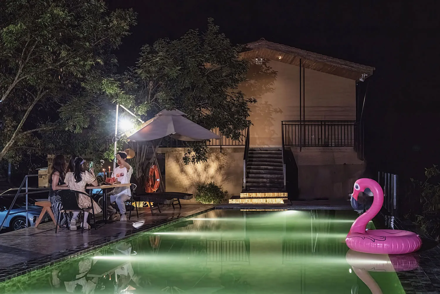 夜晚的現代泳池露台,三人圍坐在餐桌旁用餐,水下泳池燈光投射出綠色光暈,大型粉紅火鶴充氣浮圈,搭配LED照明的木質階梯與現代建築背景。