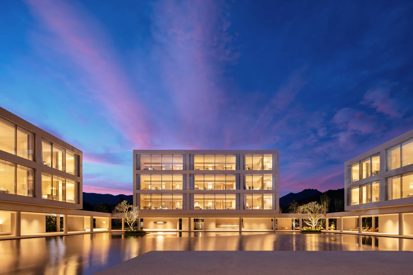 Modern multi-level concrete and glass villa complex at dusk with illuminated interiors, framed by a large reflecting pool and dramatic pink and blue sky.