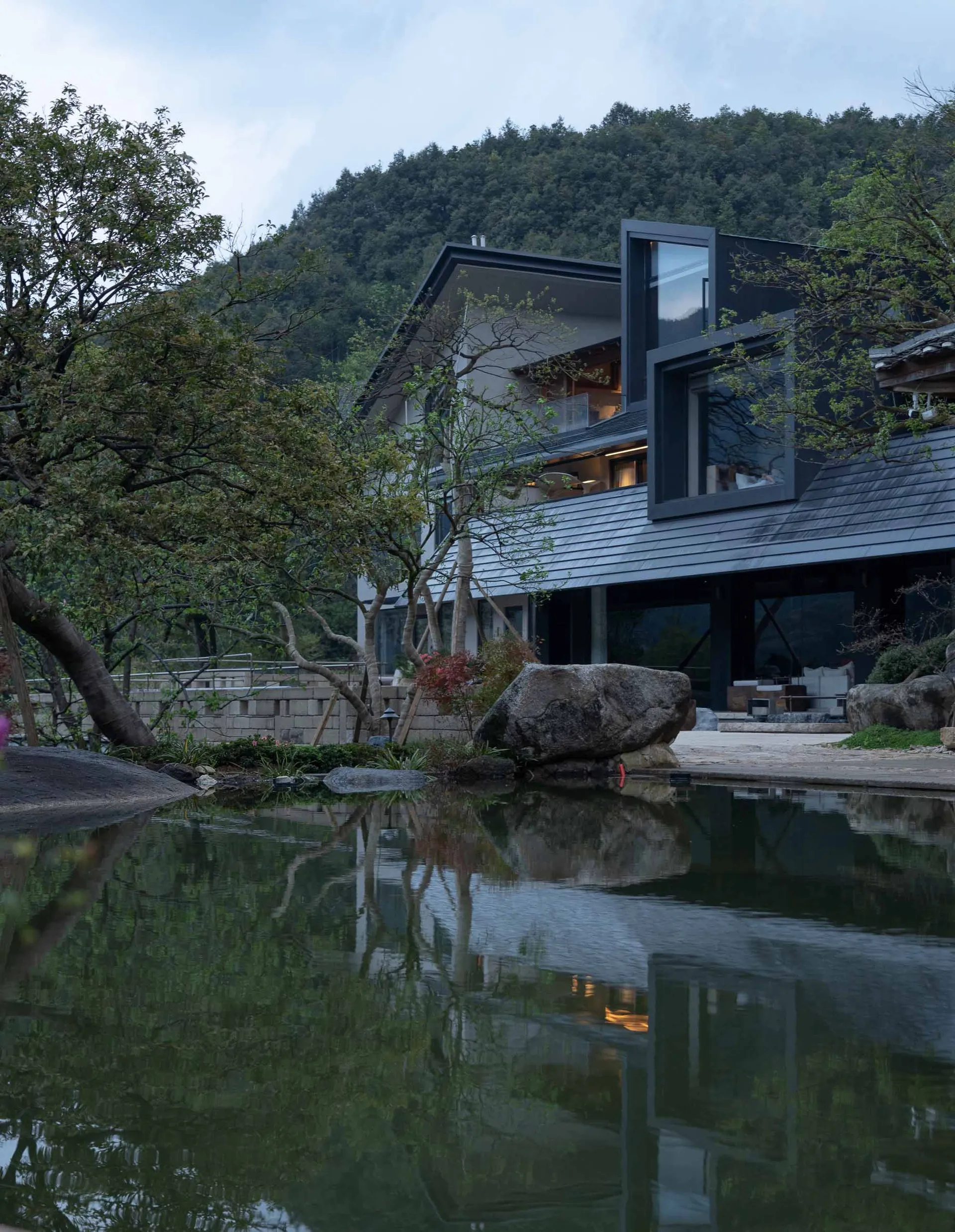 Modern minimalist villa with large glass windows and dark metal frames, surrounded by a serene reflecting pond, large natural stones, and lush trees against a forested mountain background at dusk.