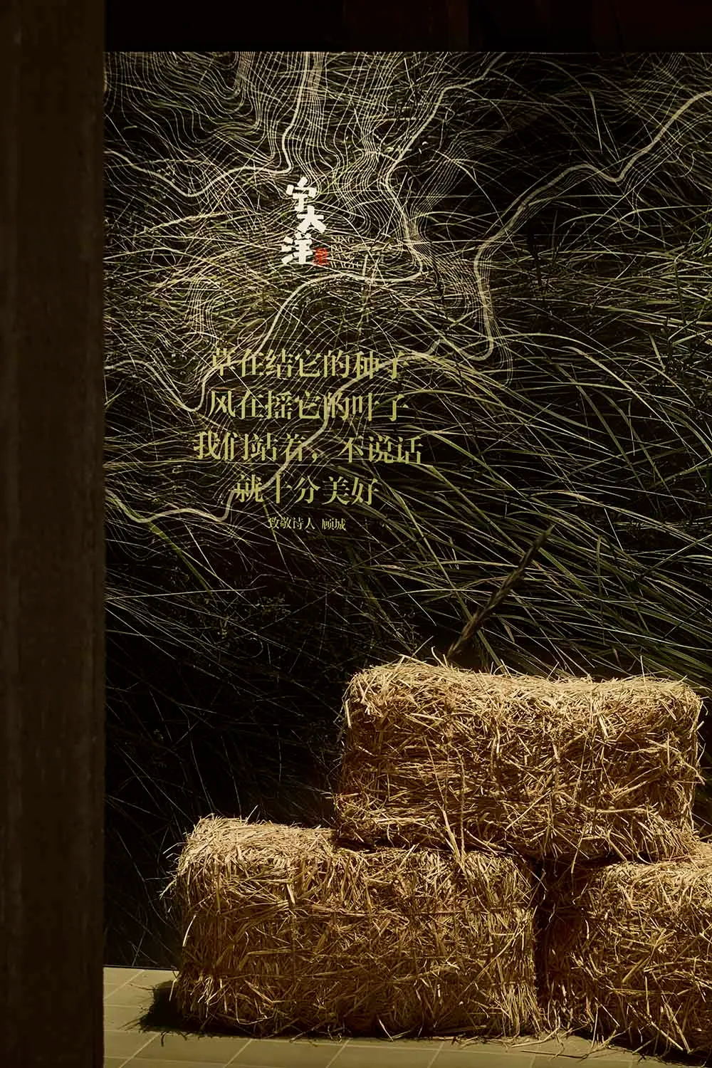 Three rectangular hay bales placed on a tiled floor in front of a dark wall featuring intricate white line art resembling grass or topography, overlaid with glowing golden Chinese poetry by poet Yu Jian.