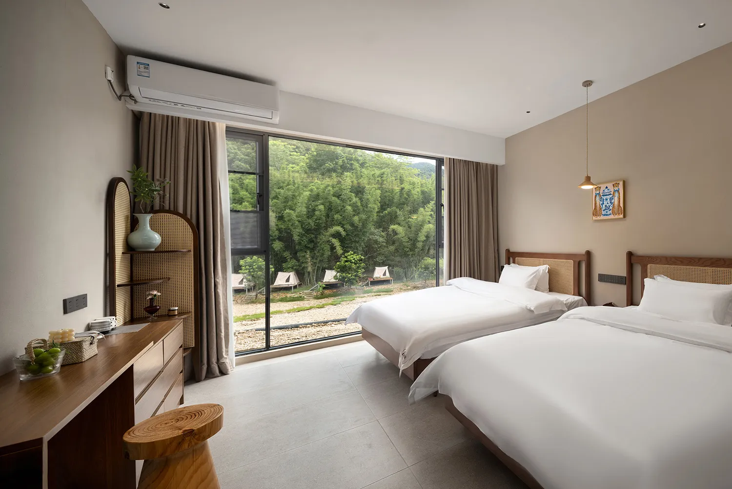 Modern minimalist twin hotel room featuring two beds with wooden and rattan headboards, a large floor-to-ceiling window overlooking a bamboo forest and outdoor tents, wooden desk with live-edge stool, and neutral beige walls.