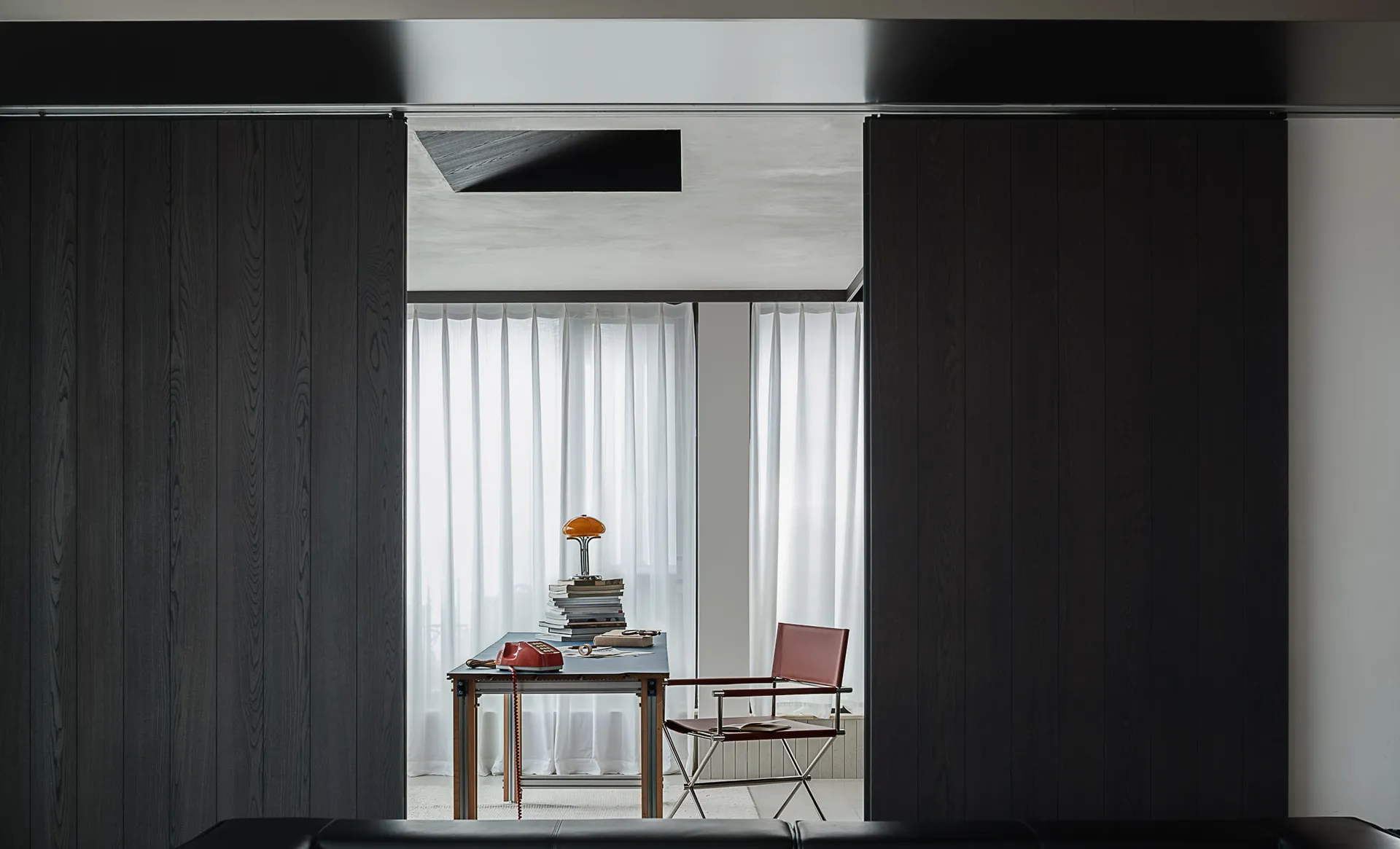 Modern minimalist home office viewed through dark wood sliding doors, featuring a blue desk with vintage red telephone, stacked books, an orange mushroom lamp, and a red leather director’s chair.