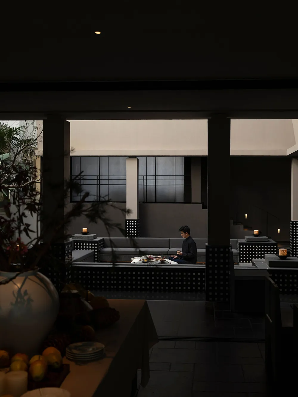 Night view of a modern minimalist concrete courtyard with built-in grey sofas, a man sitting in the sunken lounge area, warm candlelight, and dark architectural pillars.