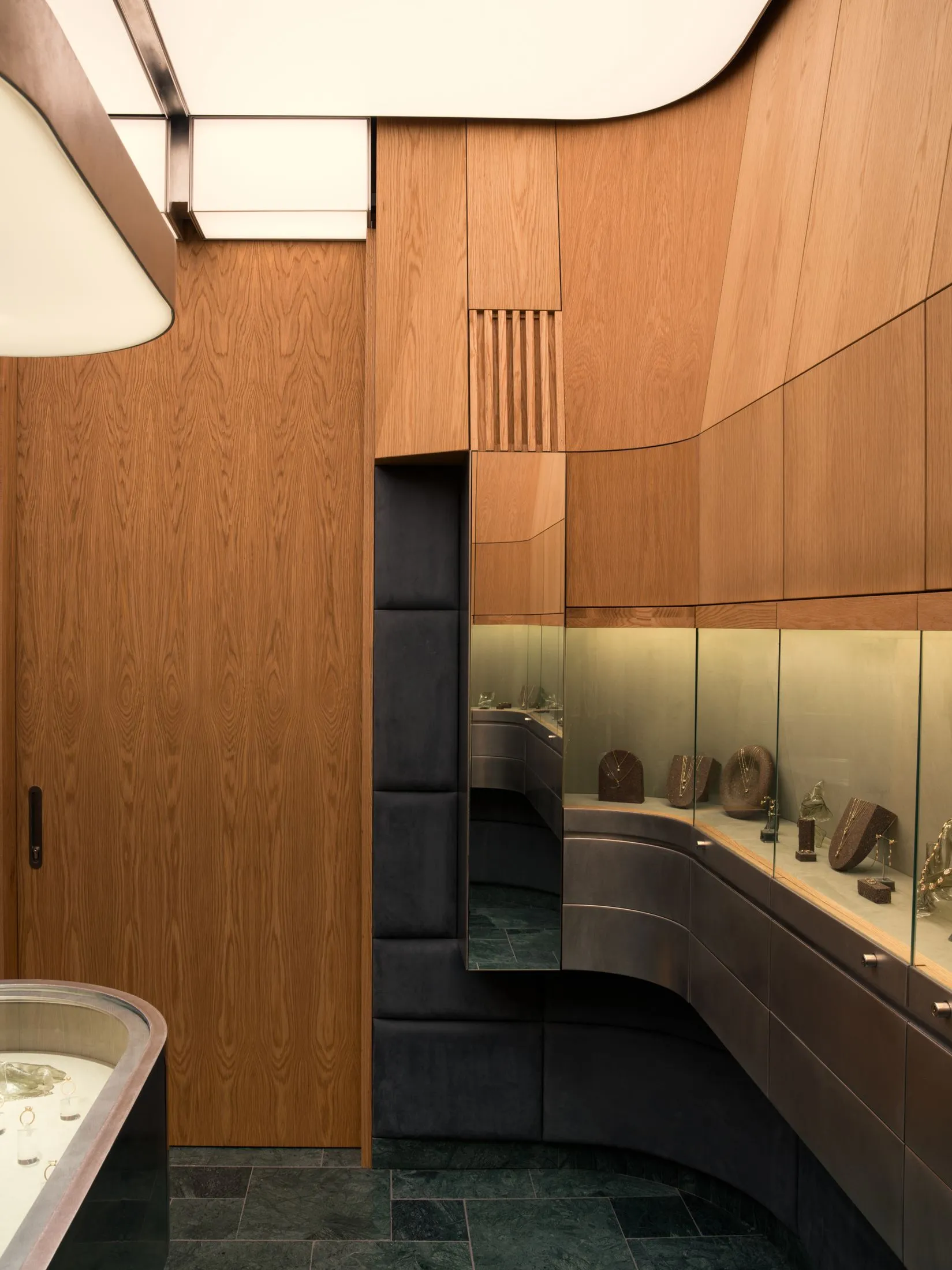 Luxury jewelry boutique interior featuring curved walnut wood paneling, dark green marble floor tiles, and illuminated glass display cases with jewelry.