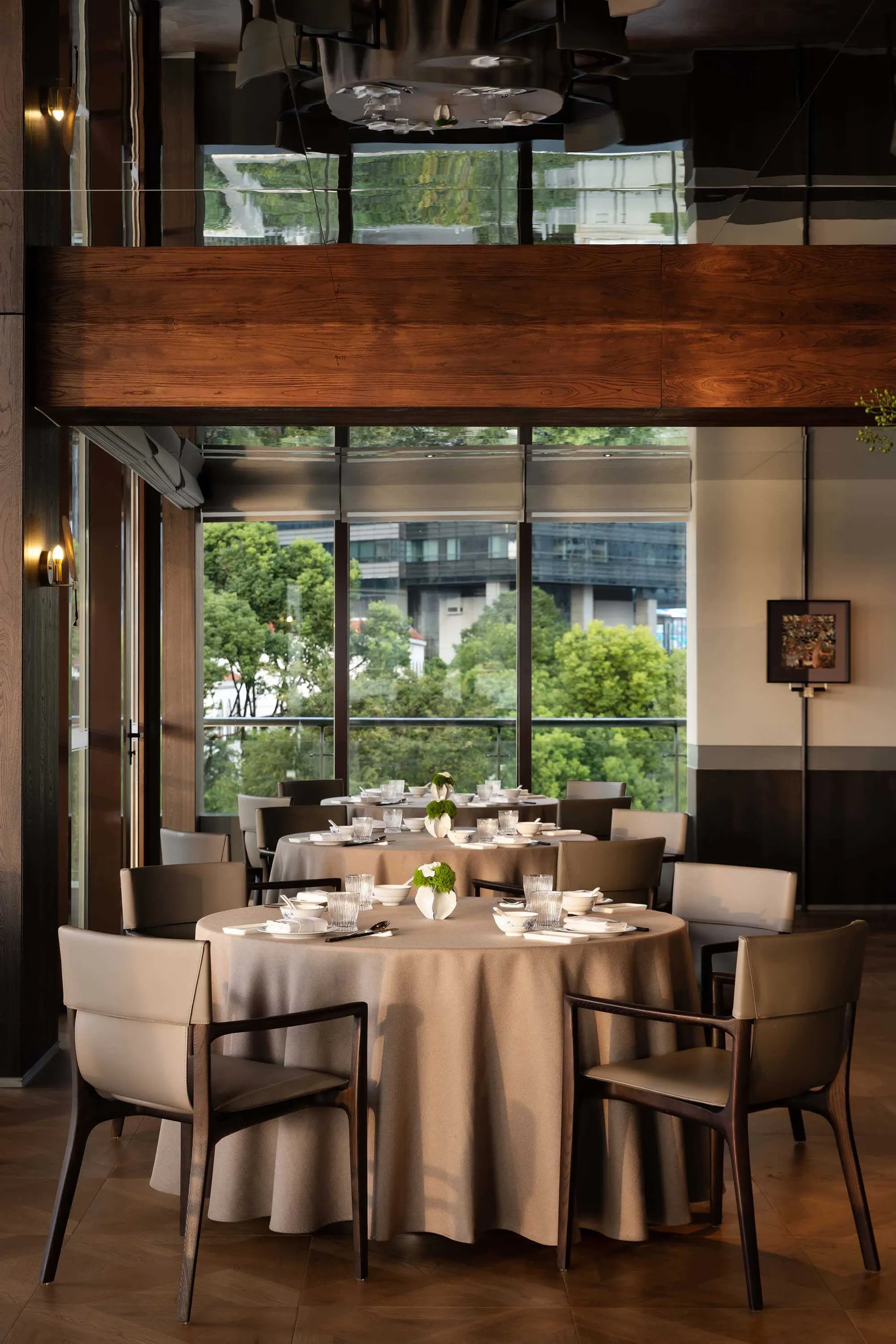 Elegant modern restaurant interior featuring round dining tables with beige tablecloths, walnut wood beams, leather-upholstered chairs, and large windows overlooking green trees and city buildings.