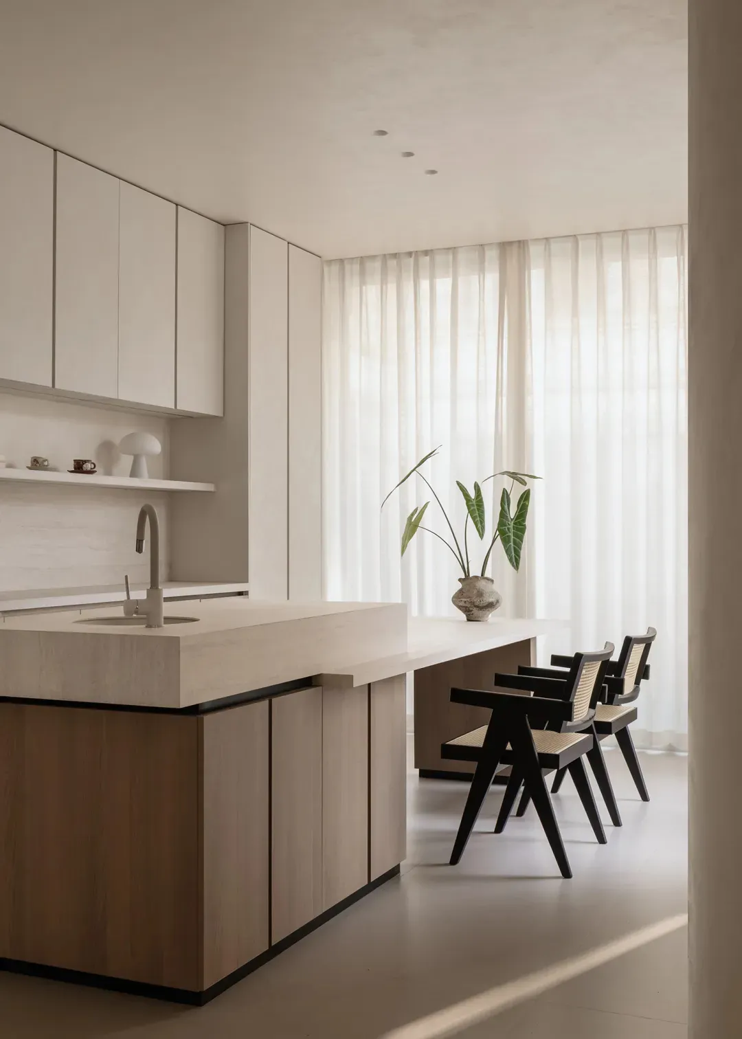 Modern minimalist kitchen featuring a large oak island with integrated sink, white upper cabinets, and three black dining chairs with cane backs placed beside a long table near a window with sheer curtains.