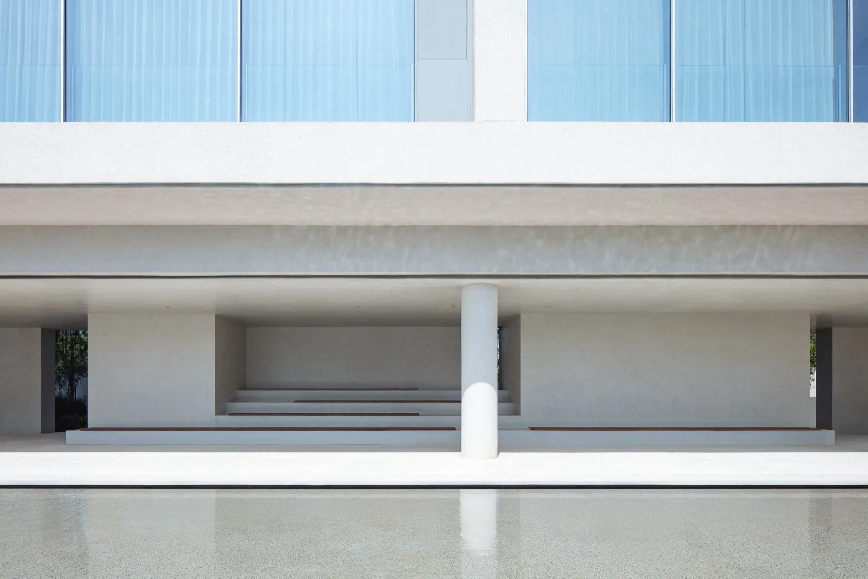 Modern minimalist concrete building facade with large glass windows, white cylindrical column, stepped seating, and a calm reflecting pool in the foreground.