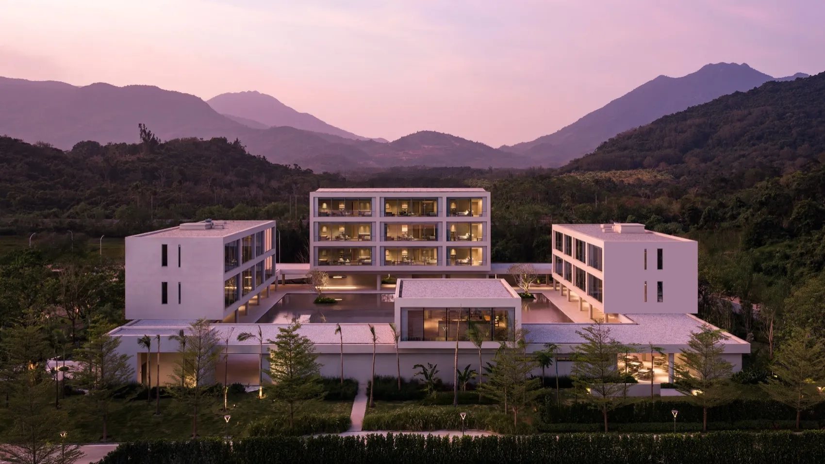 暮色中俯瞰現代混凝土度假村建築群,室內燈光通明,周圍環繞熱帶植栽與壯麗山脈。