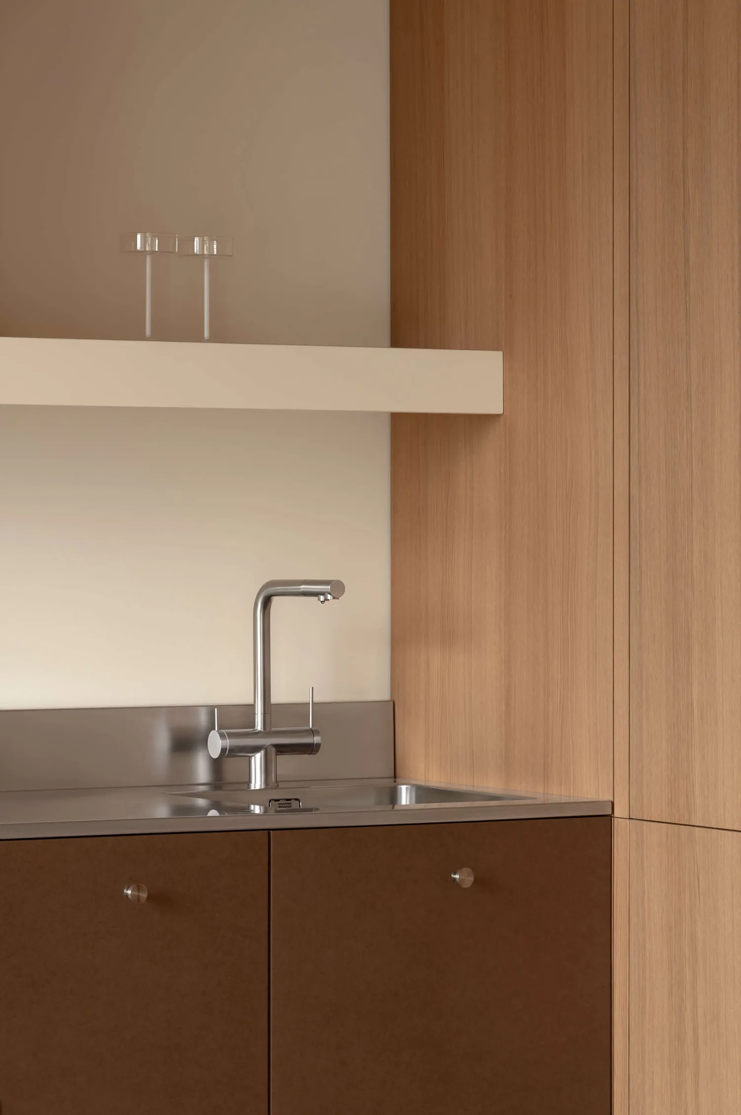 Close-up of a modern kitchen sink area with stainless steel faucet, integrated stainless steel backsplash and sink, warm wood lower cabinets with round pulls, and a floating white shelf above with two glass tumblers.
