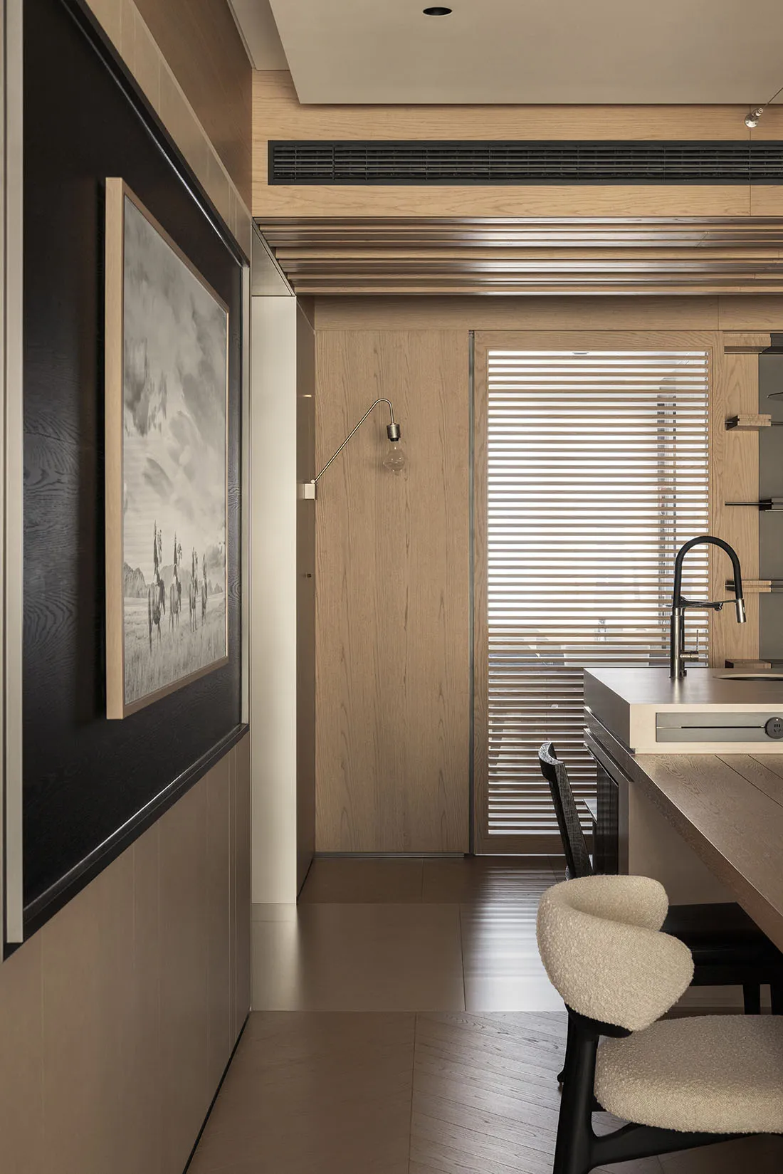 Modern minimalist kitchen featuring light oak cabinetry, black accents, boucle bar stools, and a large framed landscape photograph on a dark wood wall panel.