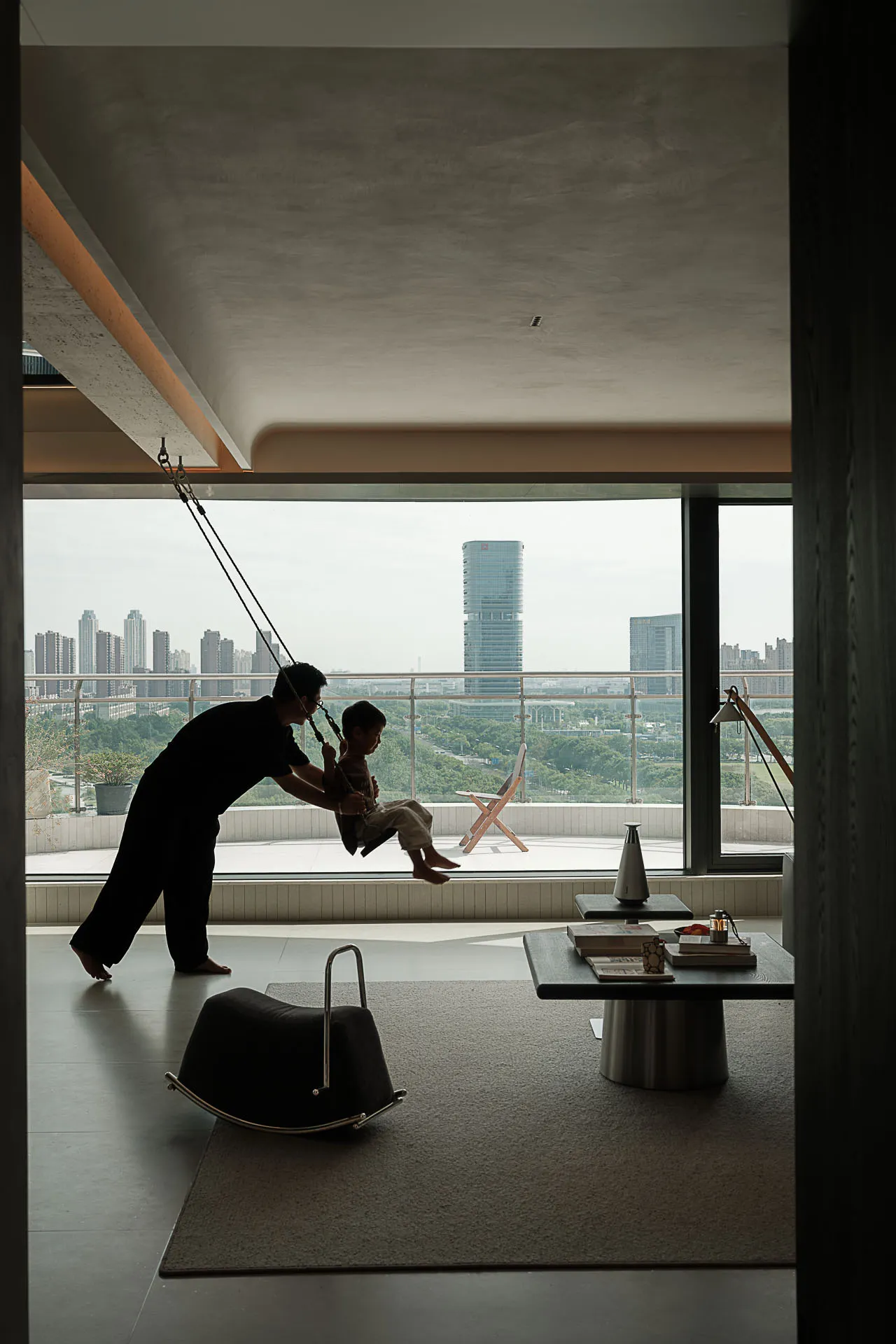 Modern minimalist living room featuring a suspended indoor swing, concrete ceiling, dark wood coffee table, and large floor-to-ceiling windows overlooking a city skyline with a father and son playing.
