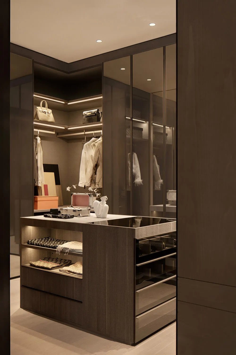Modern luxury walk-in closet with dark smoked oak cabinetry, white marble-topped central island with built-in drawers and open shelving displaying folded scarves, white ceramic vases, and luxury handbags on backlit upper shelves.