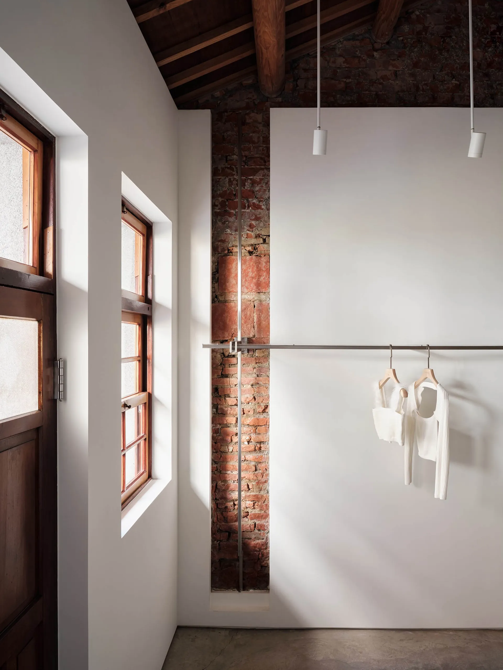 Interior corner of an industrial loft featuring an exposed red brick wall, white painted surfaces, wooden ceiling beams, a metal clothing rail with white garments, and natural light from a wooden window.