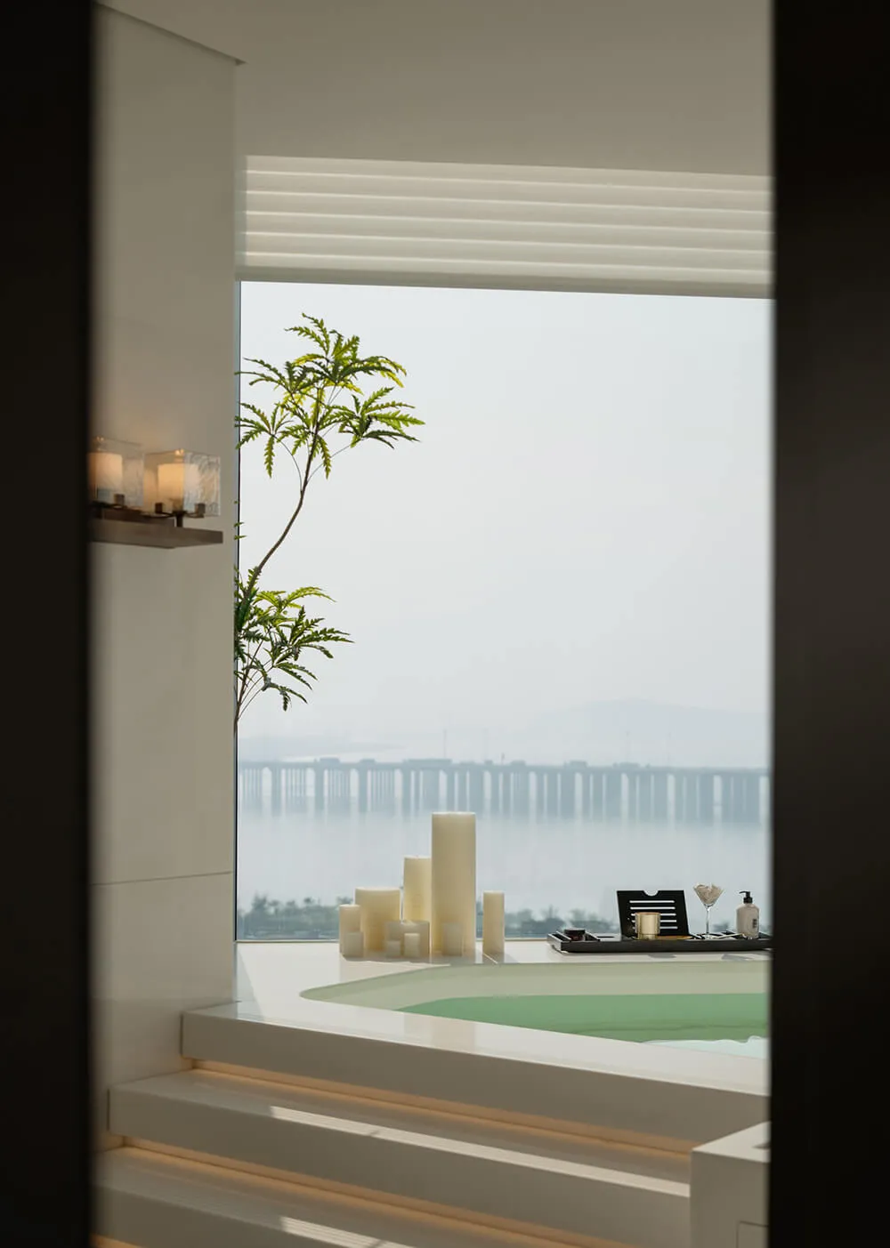 Modern minimalist bathroom featuring a built-in green marble bathtub with integrated steps, white marble walls, large ocean view window, potted plant, and decorative candles.