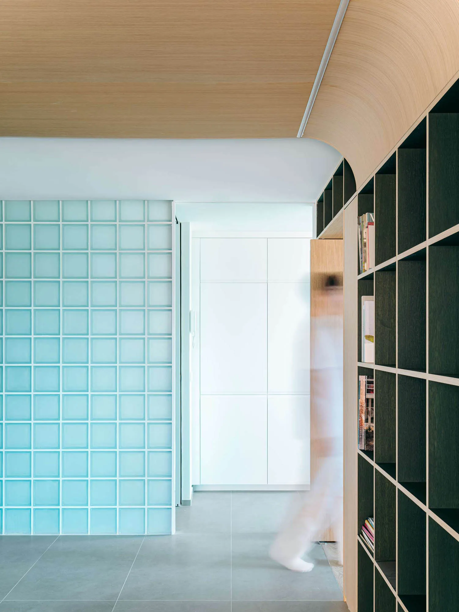 Modern minimalist hallway featuring a curved light-wood ceiling, translucent turquoise glass block wall, dark green built-in bookshelf, and grey tiled floor with a person walking by.