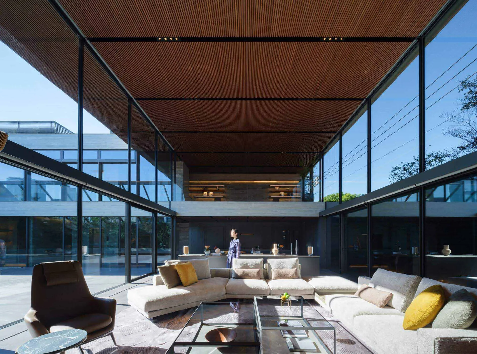 Modern double-height living room featuring a large L-shaped sectional sofa, brown leather armchair, glass coffee tables, and dramatic wood slat ceiling with full-height glass walls overlooking a courtyard.