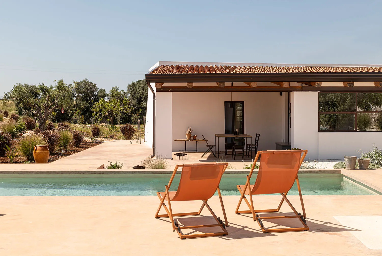 Two wooden orange deck chairs placed on a light-colored concrete terrace next to a swimming pool, with a white Mediterranean-style house featuring a terracotta tile roof and outdoor dining area in the background.